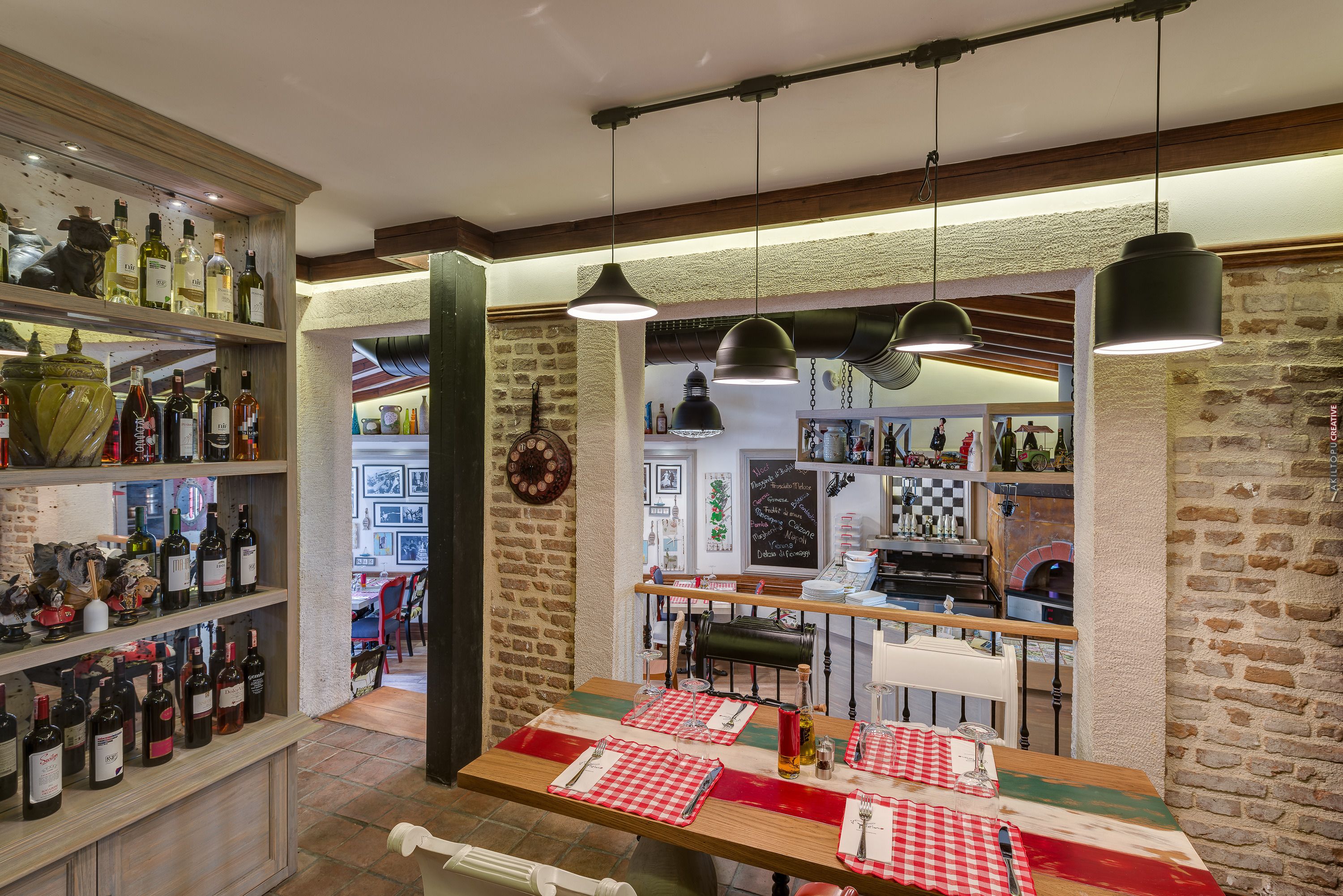 Ein Restaurant mit Backsteinwand, Weinfleschen auf Holzböden und einem Tisch mit rot-weiß kariertem Tuch, dazu hängende Pendelleuchten.