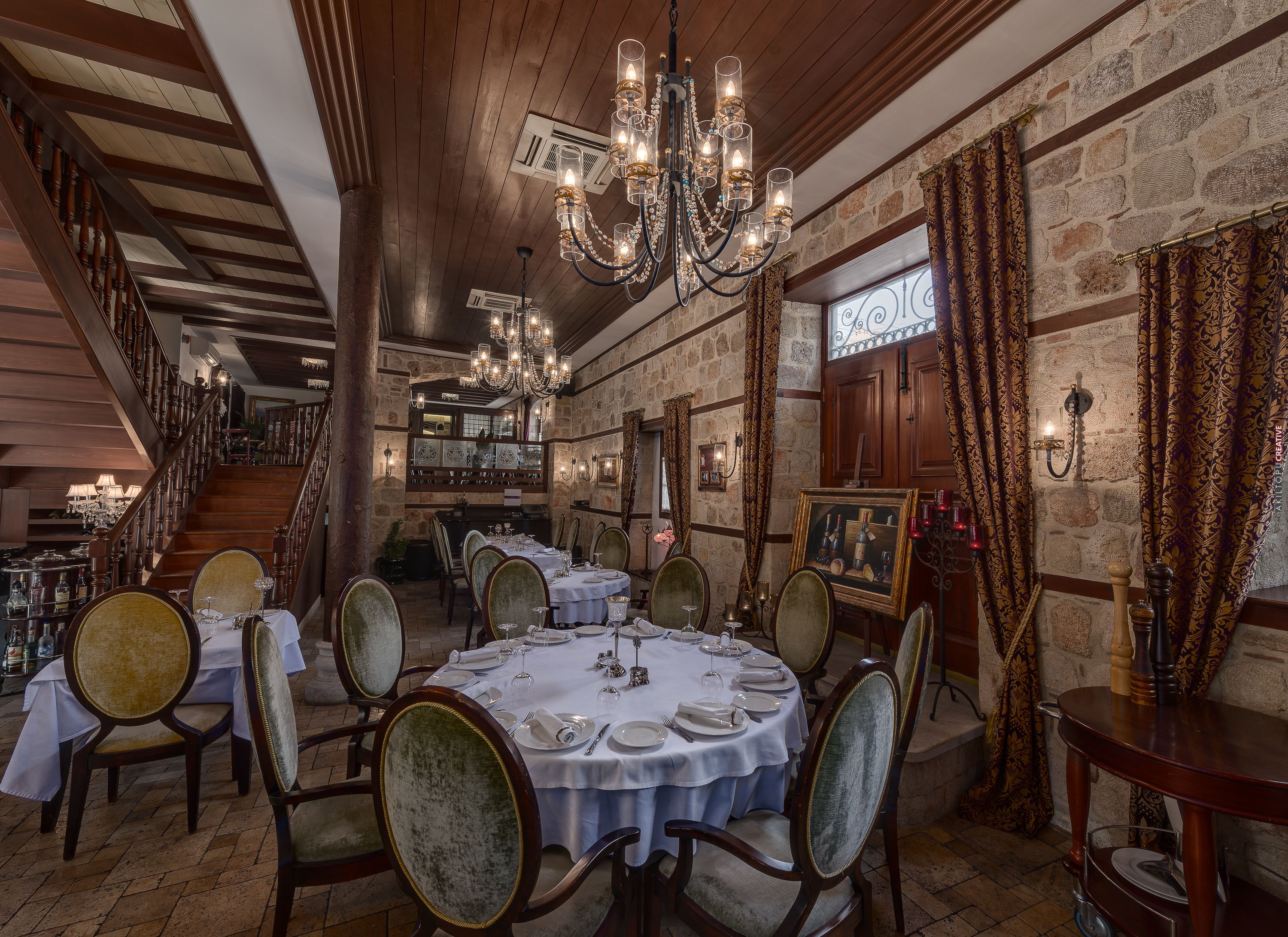 A dining area with round tables set for four, wooden chairs, a chandelier, stone walls, and a staircase.