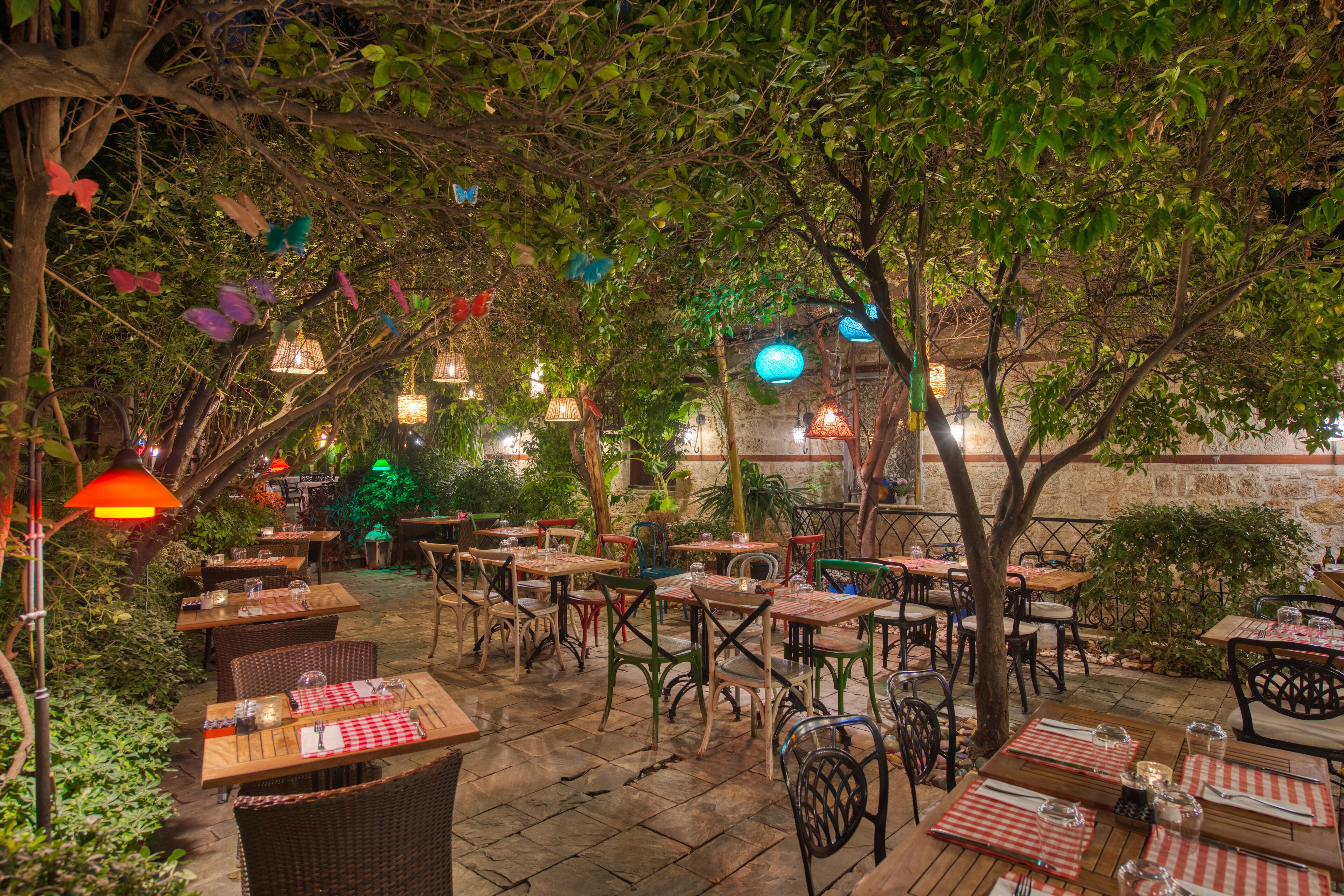 An outdoor dining area with tables and chairs, surrounded by trees and hanging lanterns.