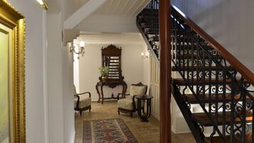 Una gran escalera con barandillas negras ornamentales que conduce a un rellano. Una alfombra estampada y dos sillones tapizados se encuentran en el rellano, junto con una consola de madera y un espejo encima.