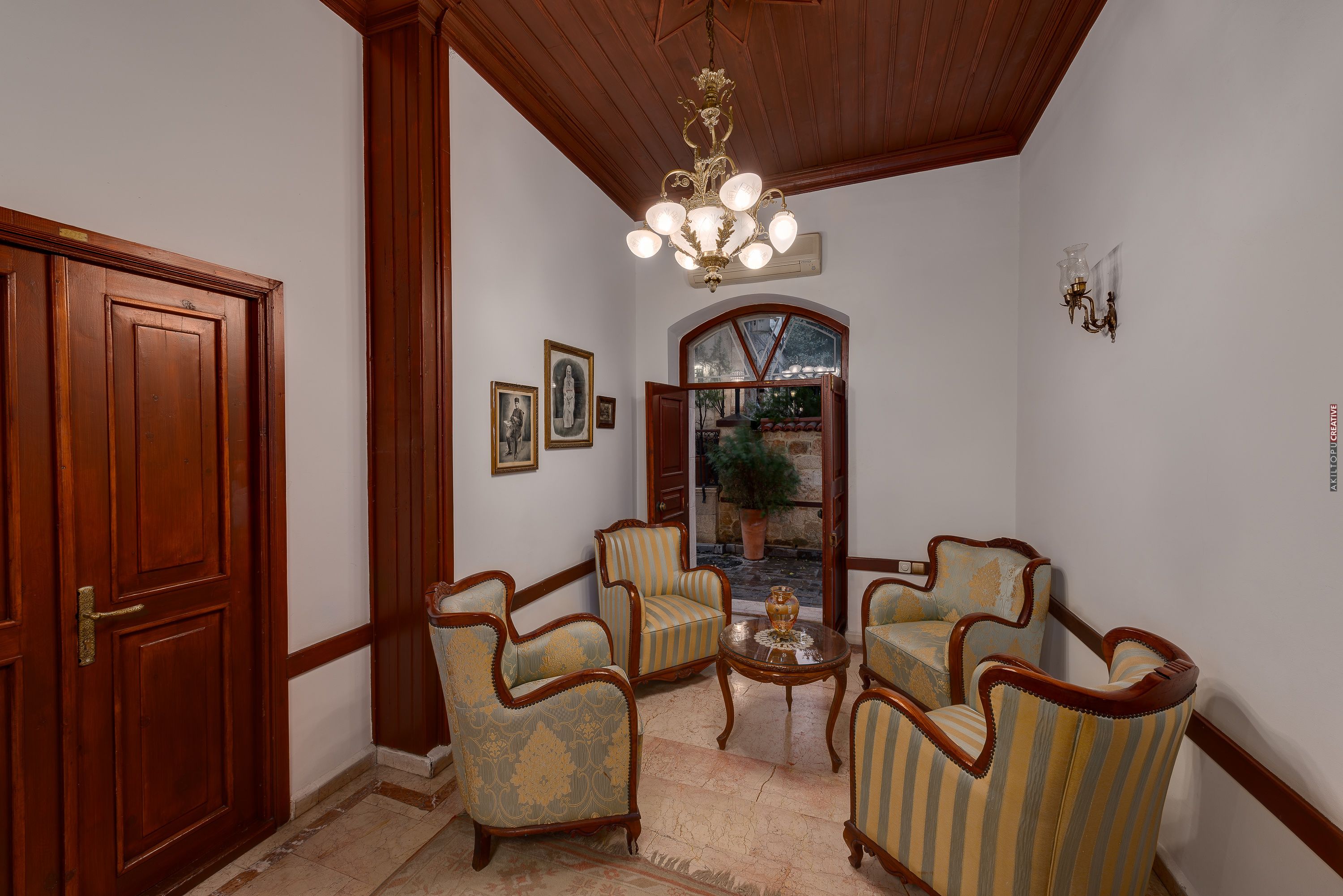 A room with wooden paneling, a chandelier, four upholstered chairs, a small table, and framed pictures on the wall.