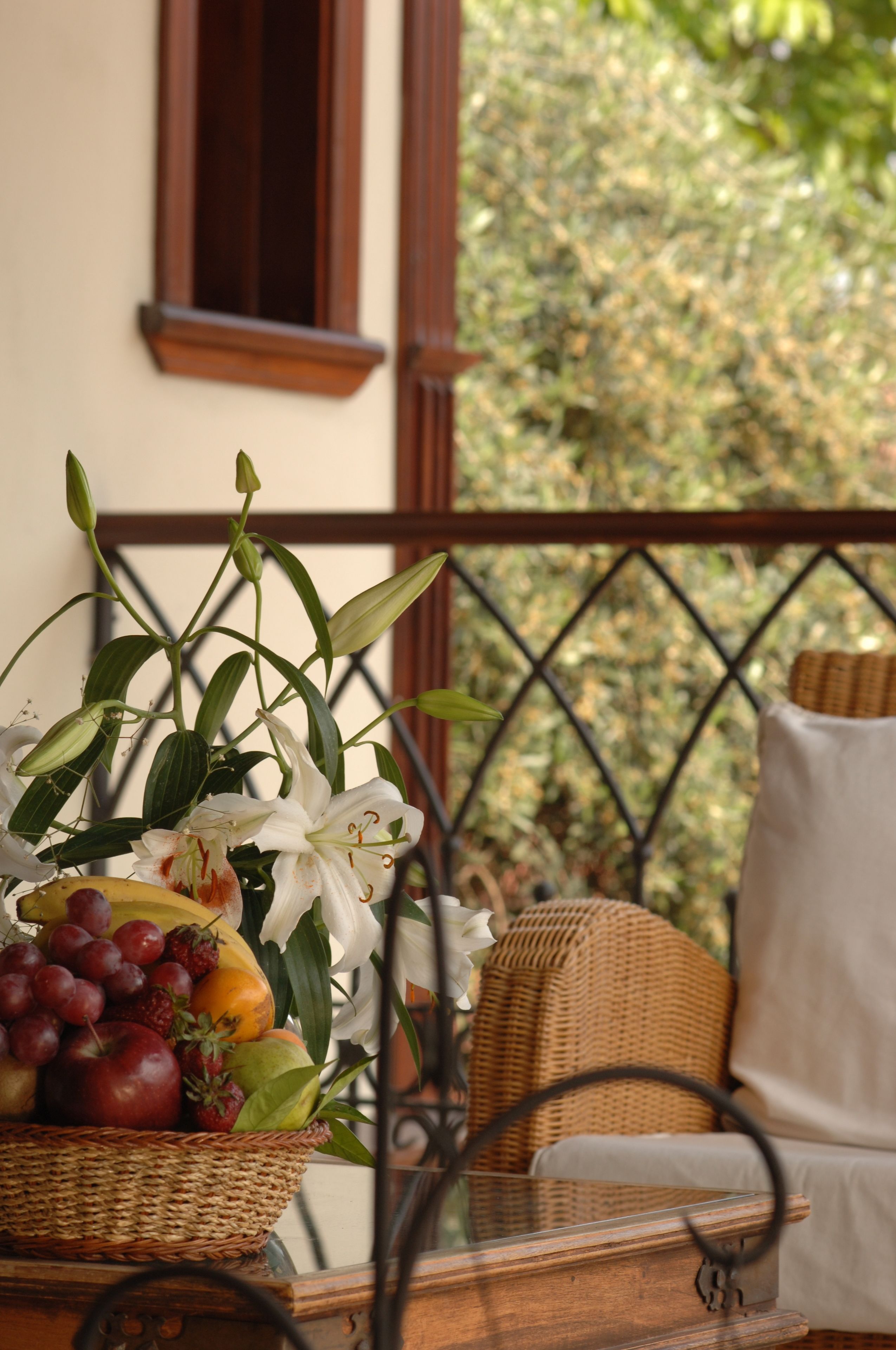 A wicker chair with a cushion, a wicker basket with fruits and flowers, and a view of greenery through a metal railing.