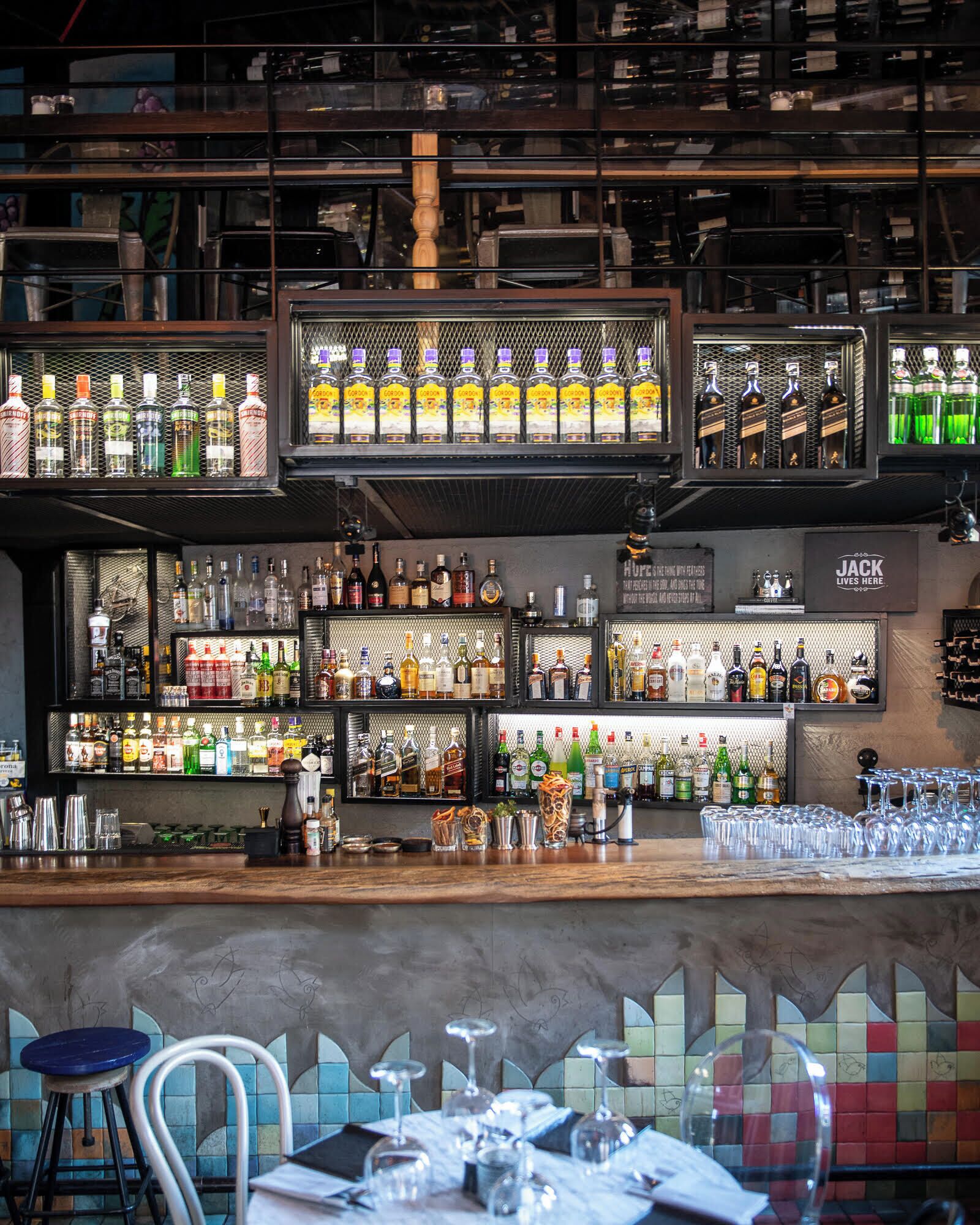 A bar area with multiple shelves of liquor bottles, a Jack Daniels sign, and a tiled backsplash.