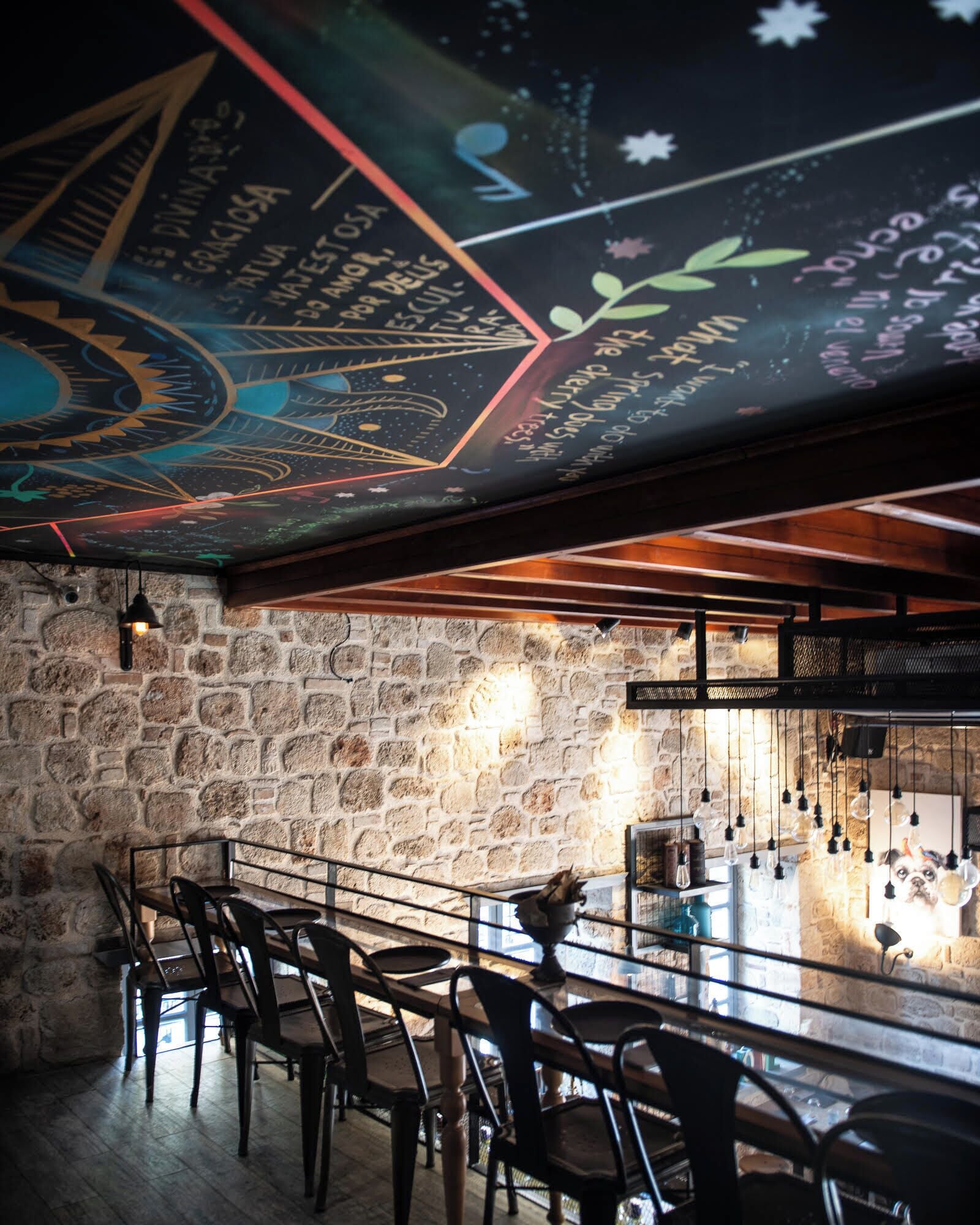 A bar area with a stone wall, wooden ceiling, and a mural. There are bar stools and a counter with a hanging light fixture.