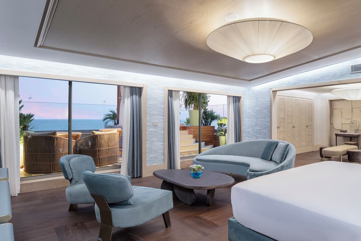 Ein geräumiges Hotelzimmer mit einem großen Bett, zwei Sesseln, einem Couchtisch und Meerblick durch große Fenster.