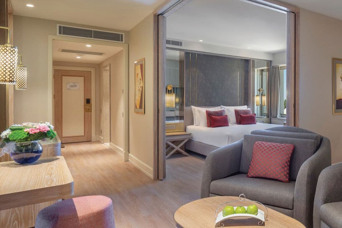 Ein modernes Hotelzimmer mit einem Bett, zwei Sesseln, einem kleinen Tisch und Blick auf die Stadt durch große Fenster.
