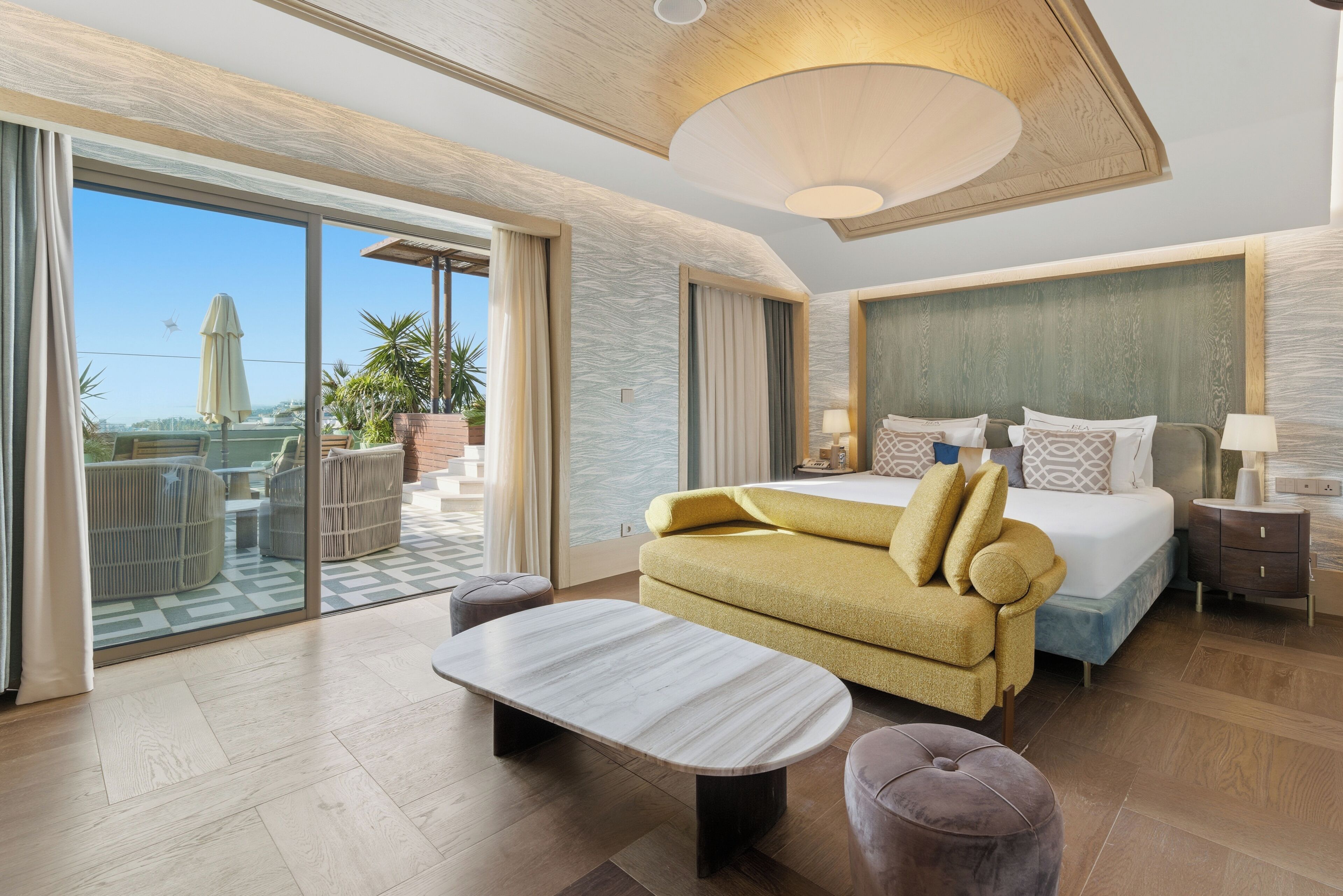 A modern hotel room with a large bed, a sofa, a dining table, and a balcony with outdoor seating.