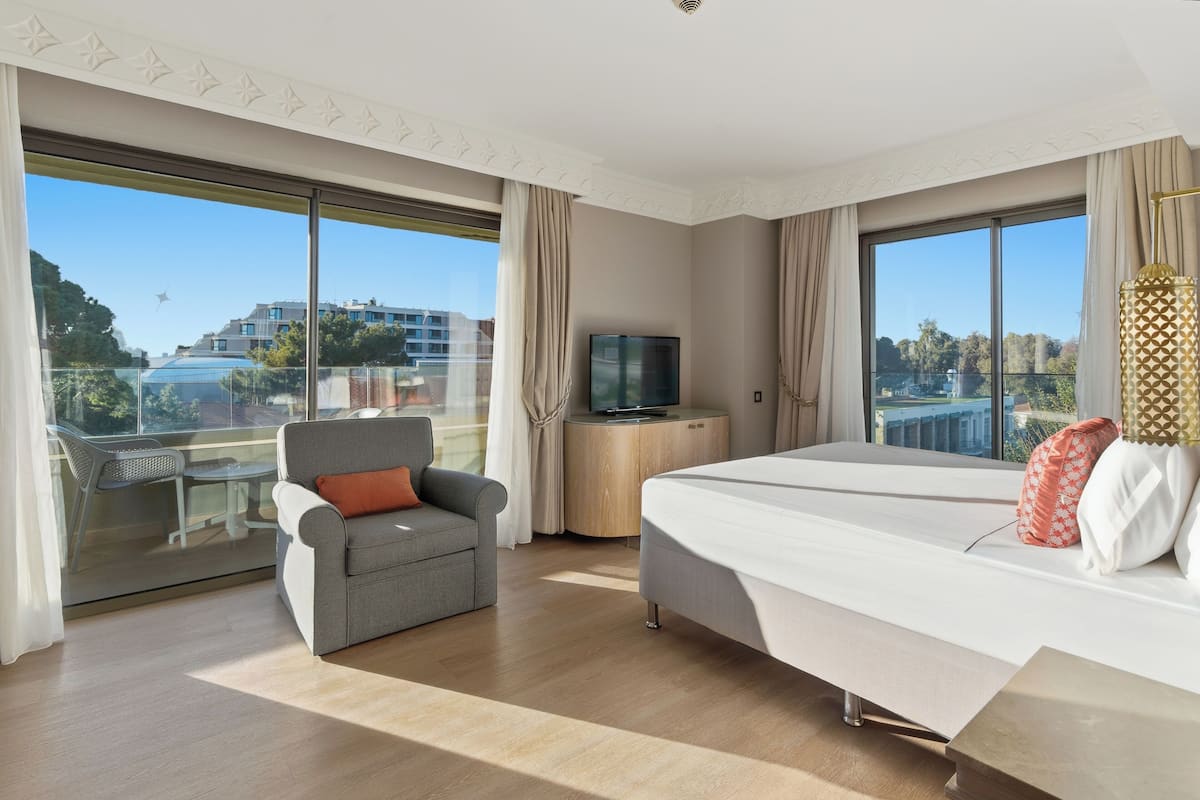 Ein Hotelzimmer mit einem großen Bett, einem grauen Sessel, einem Fernseher auf einem Holznachttisch und einem Balkon mit Blick auf Häuser und Bäume.