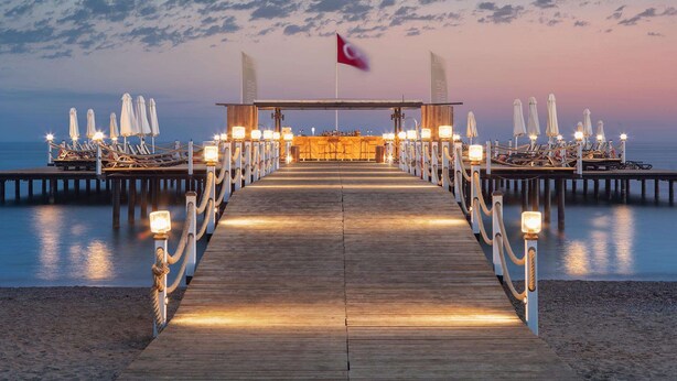 Eine Holzstege führt zu einer Strandbar mit Sonnenschirmen und Liegestühlen.