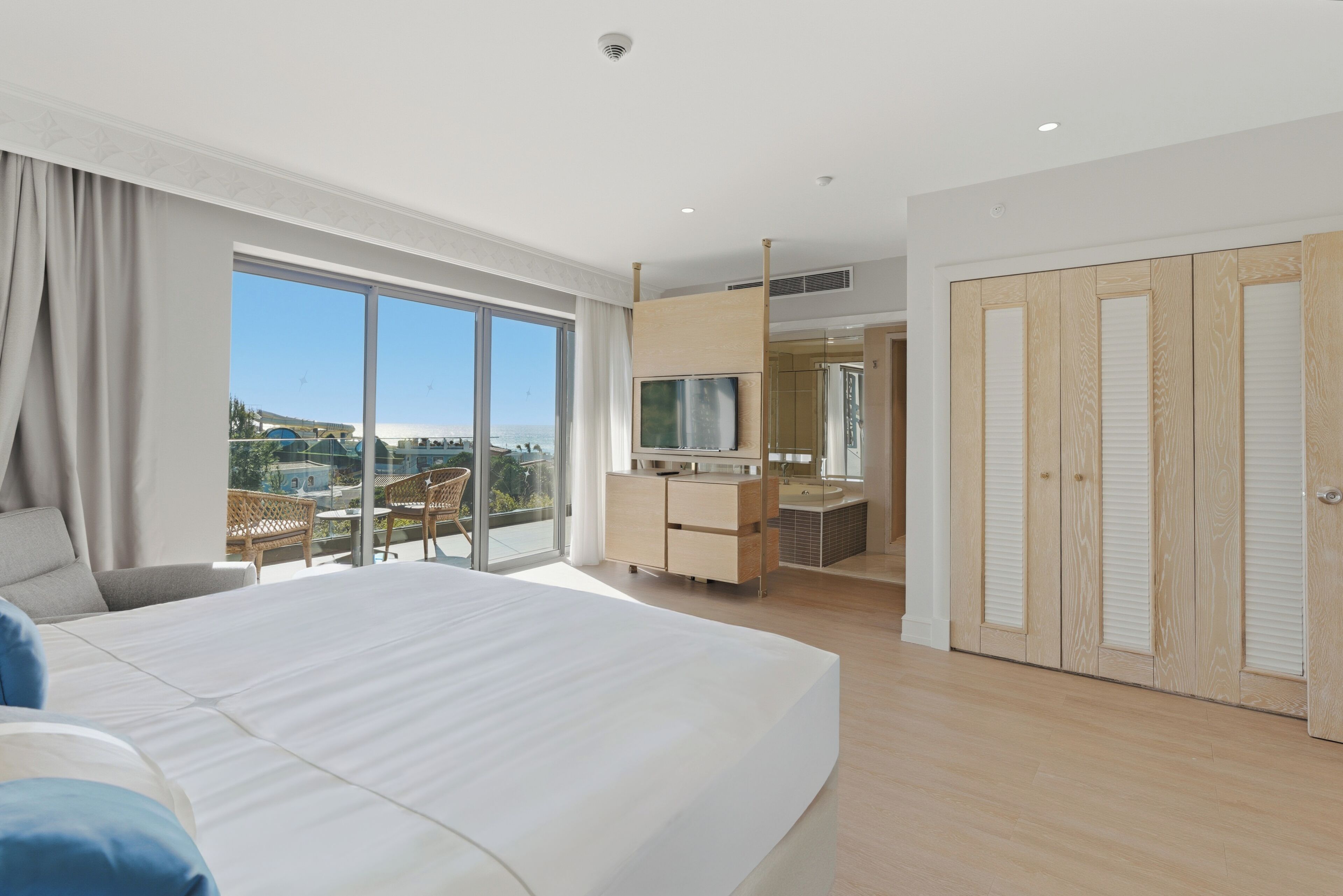 A modern hotel room with a large bed, a desk with a TV, and a balcony with a view.