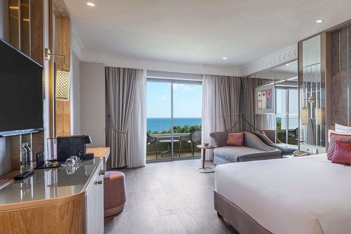 Ein Hotelzimmer mit einem großen Bett, einer Sitzecke inklusive Sofa und Sesseln, einem Balkon mit Aussicht sowie einem Fernseher auf der linken Seite.