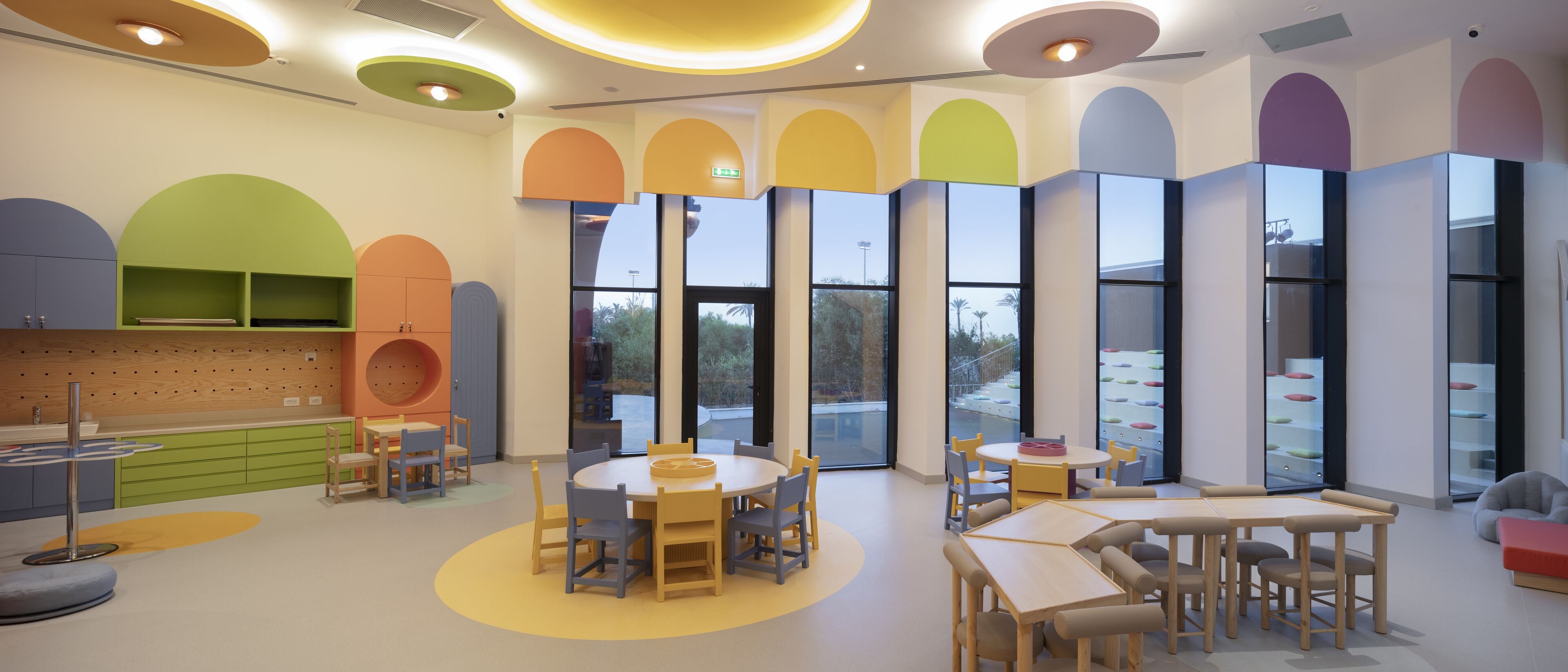 Un área de juegos infantiles bien iluminada con mesas y sillas redondas, ventanas amplias y coloridos arcos en las paredes.