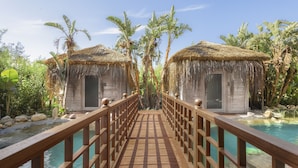 A wooden bridge leading to two thatched-roof huts by a pool, surrounded by palm trees. - ELA Excellence Resort Belek - All Inclusive (Serik)