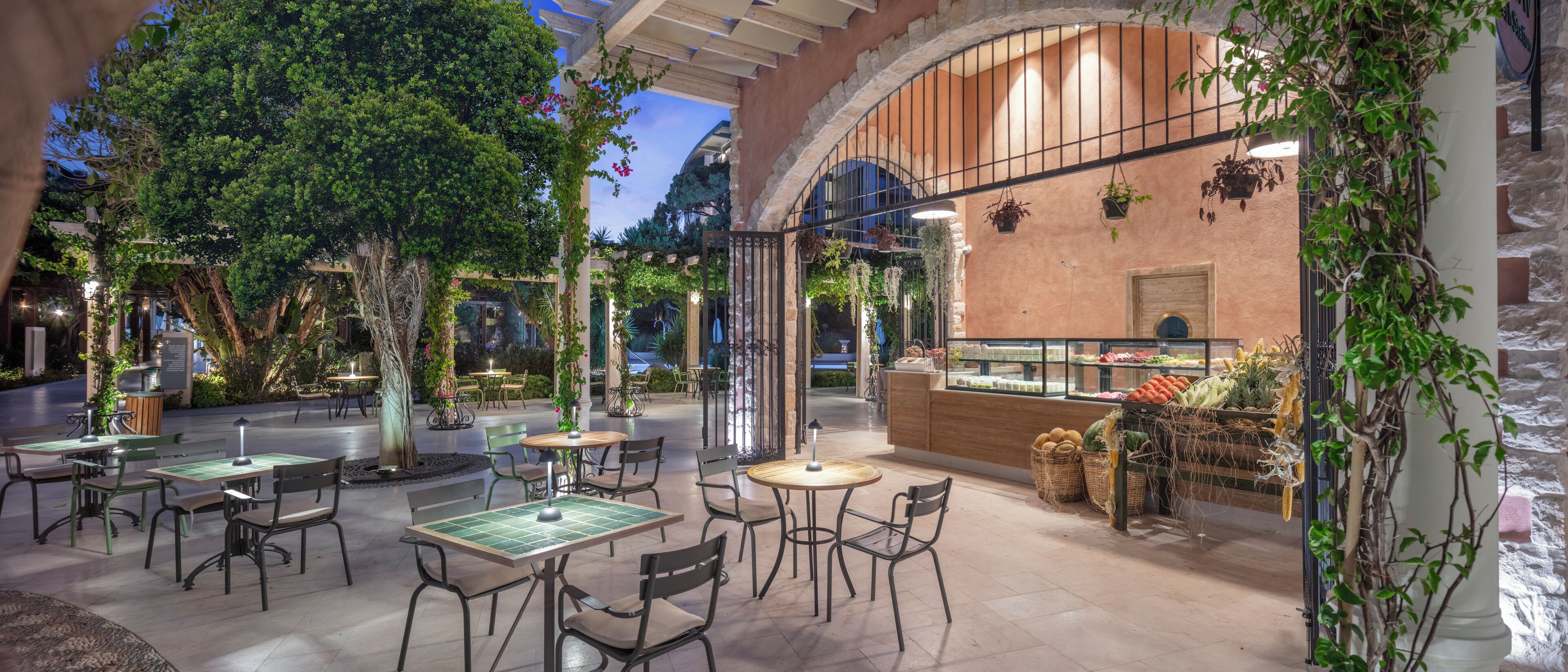 An outdoor café area with tables and chairs, surrounded by greenery and a brick wall.