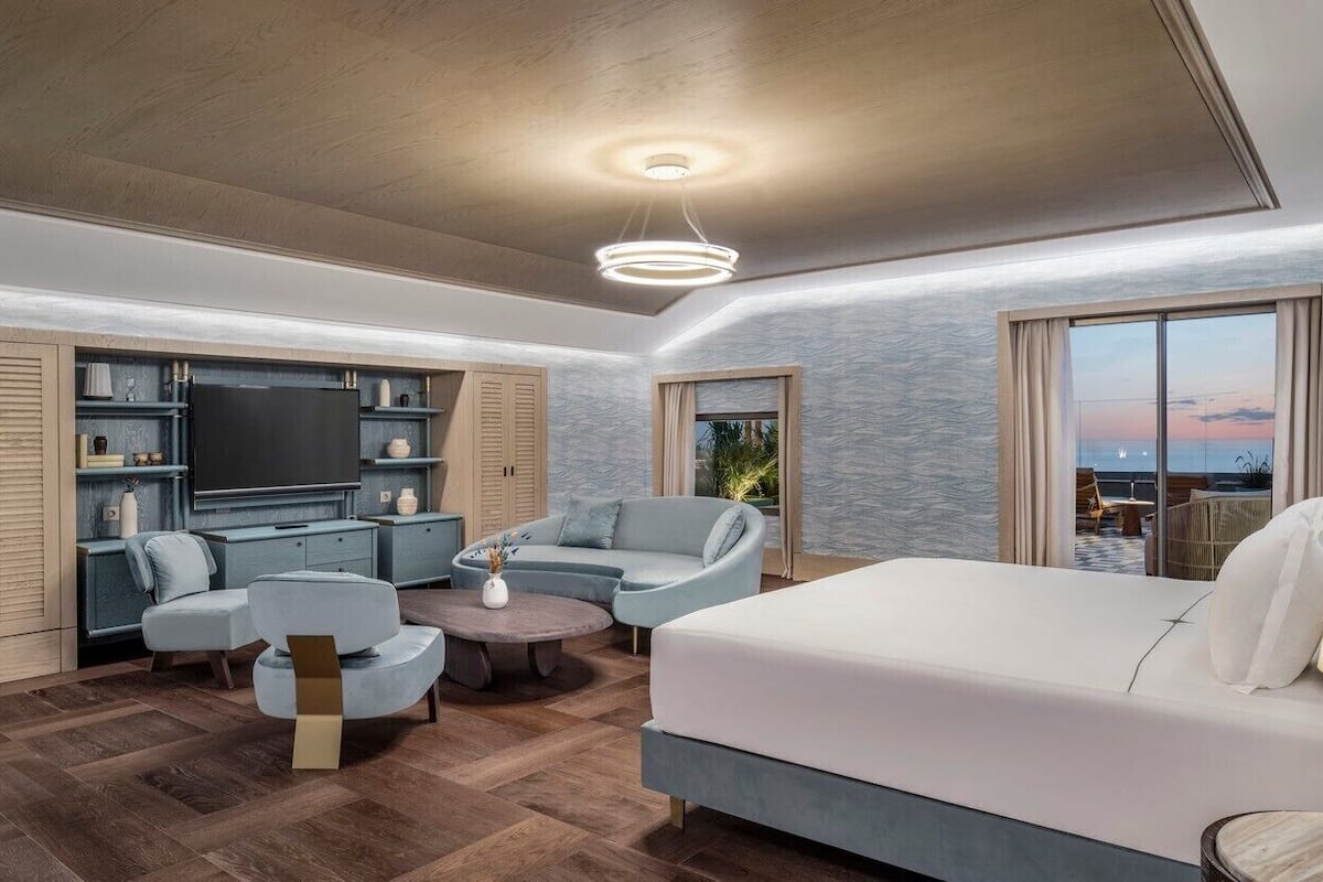 Ein modernes Hotelzimmer mit einem großen Bett, einer Sitzecke mit Sofa und Sesseln, einem Fernseher und einem Balkon mit Blick.