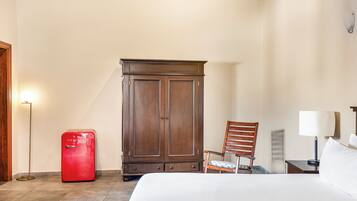 Habitación de hotel con cama, armario de madera, refrigerador rojo, una silla, mesita de noche con lámpara y una lámpara de pie.