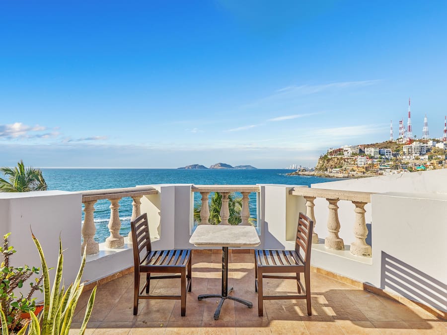 A balcony with two wooden chairs and a small table, overlooking a coastal city with buildings and a clear blue sky.