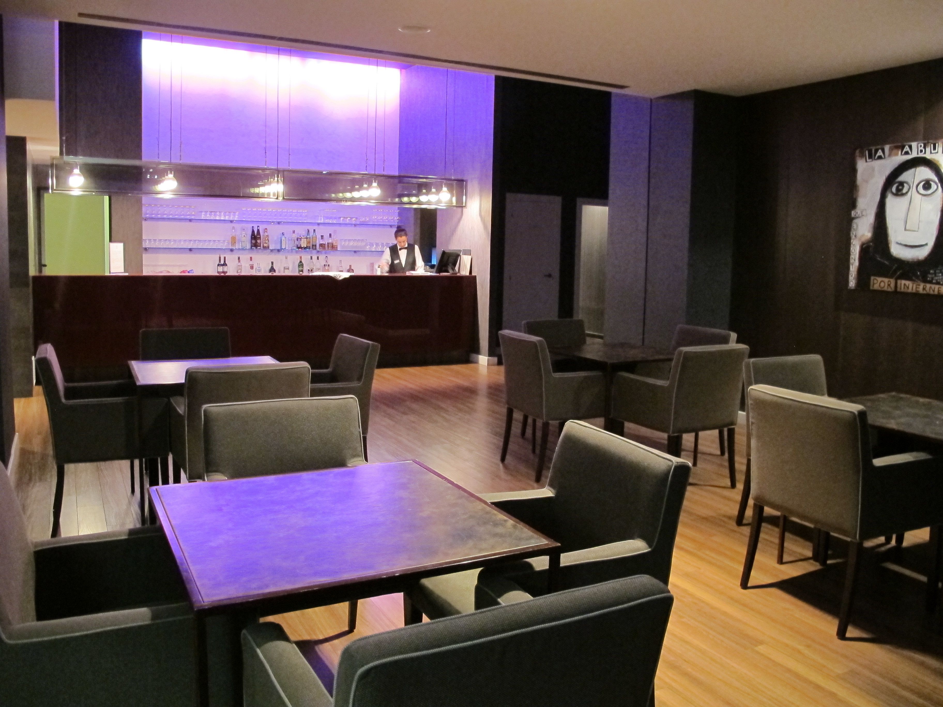 Un restaurante con zona de bar, varias mesas y sillas, y una pared con un cuadro.