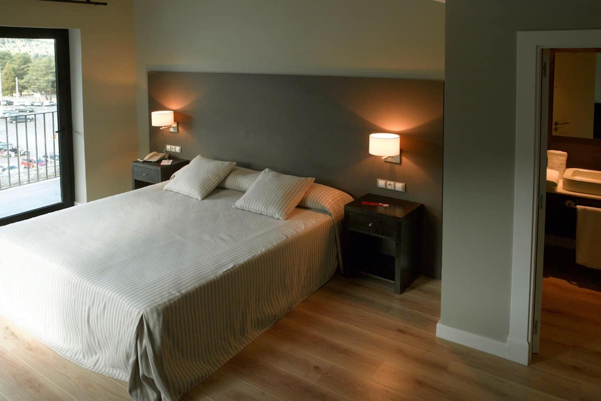 Un dormitorio con una cama grande, dos mesitas de noche con lámparas, suelo de madera y vistas al exterior a través de una puerta.
