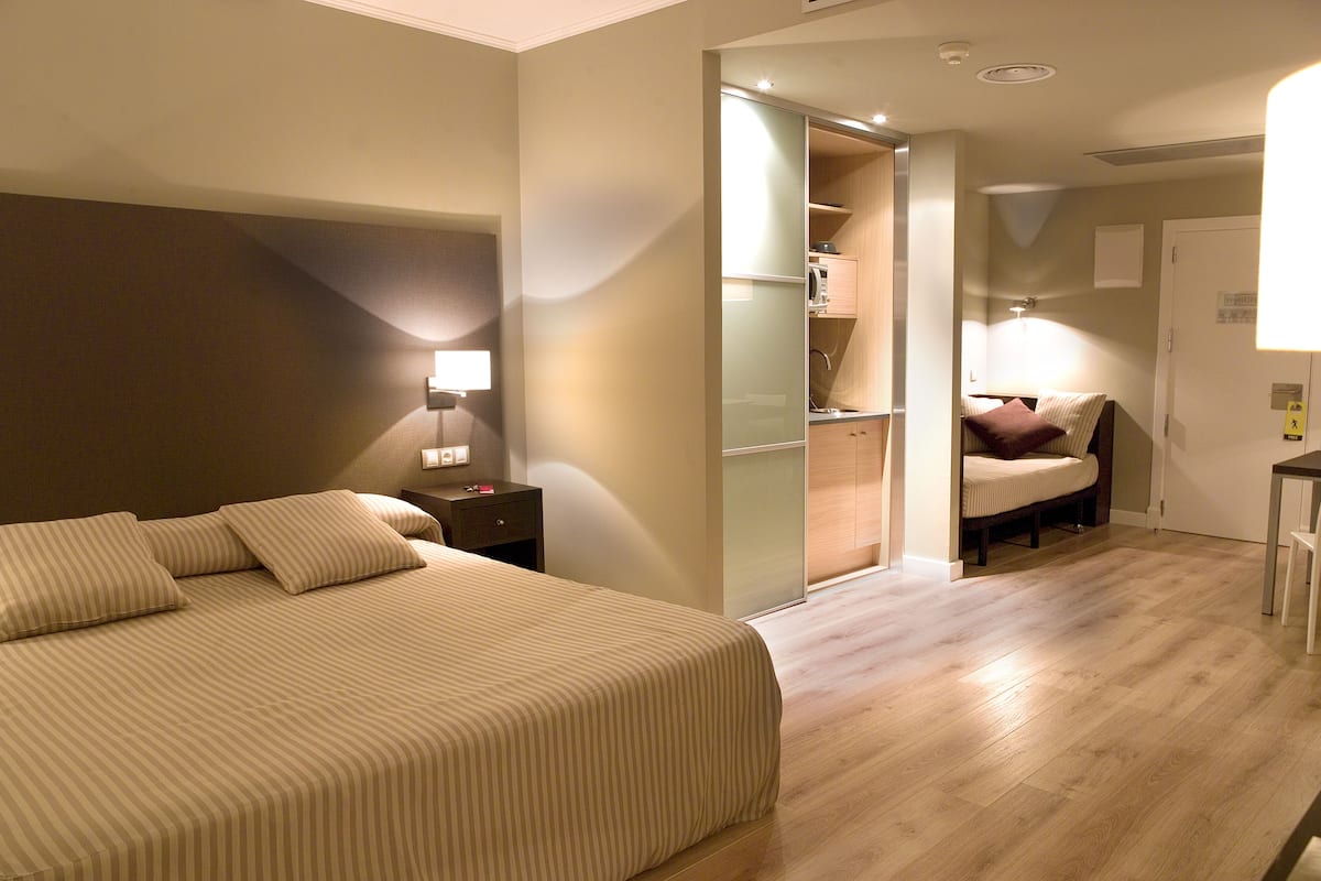 Una habitación de hotel con una cama grande, una mesita de noche, un armario empotrado y una pequeña cocina americana.