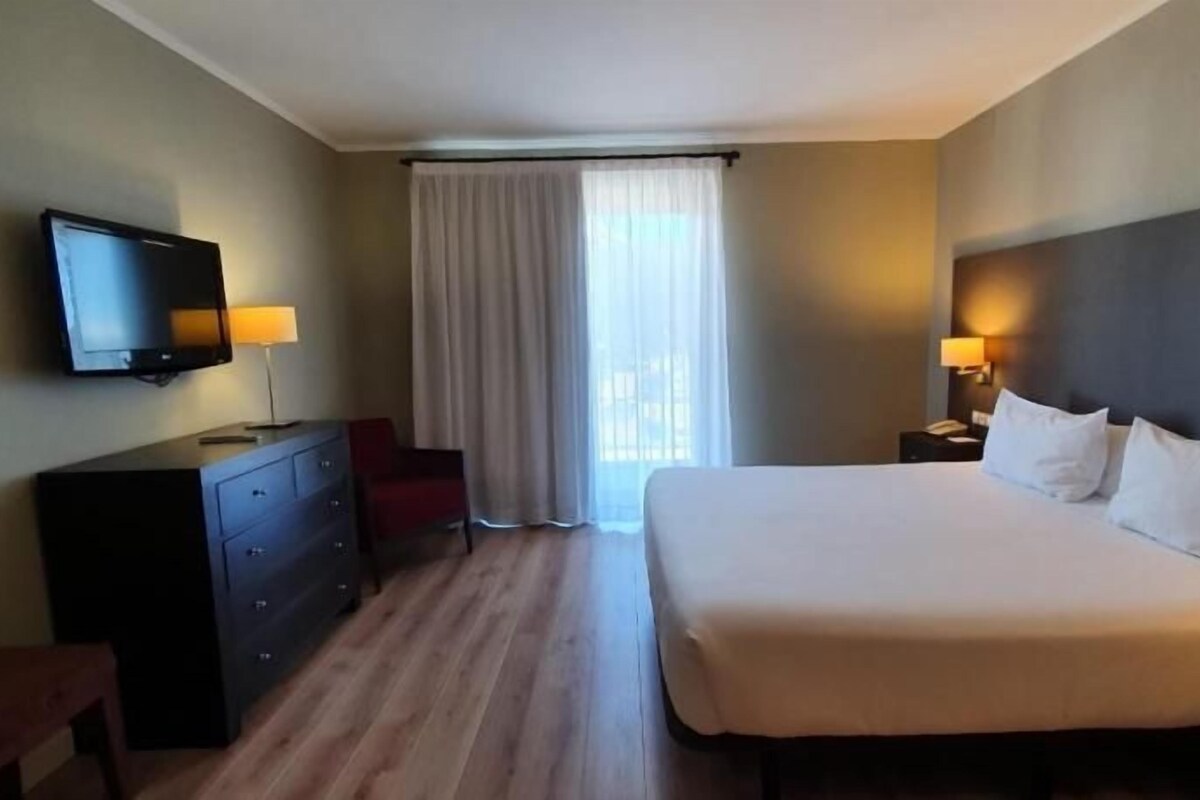 Una habitación de hotel con cama, televisión, escritorio, silla y ventana con cortinas.