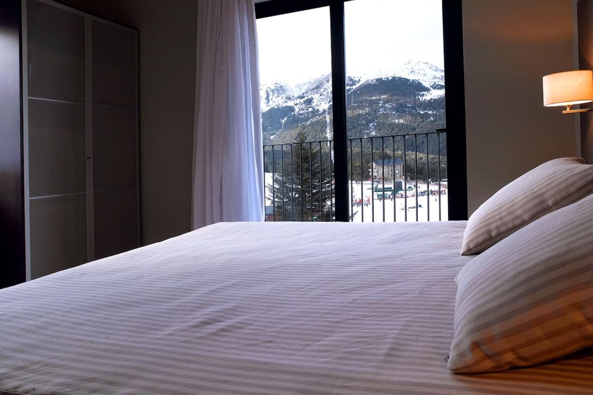 Habitación de hotel con una cama grande, un armario, un ventanal con vista a la montaña y una mesita de noche con lámpara.