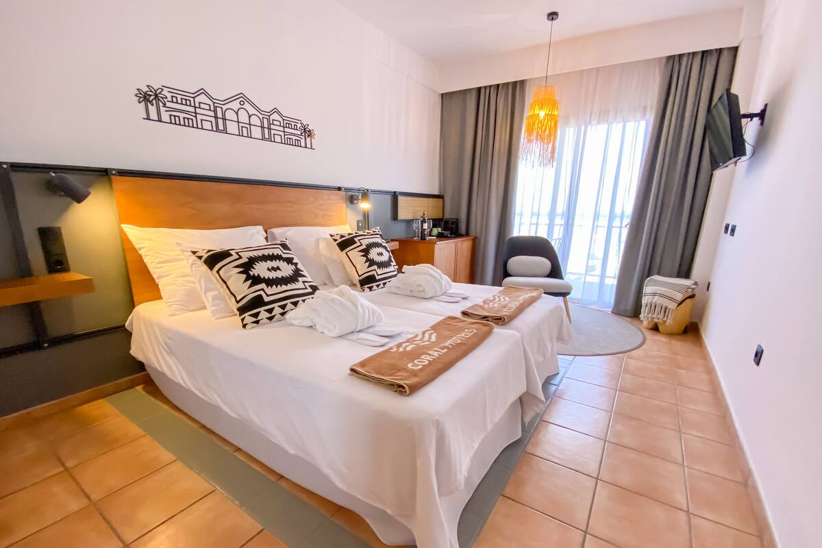 A hotel room with a large bed, two bedside tables, a desk, a chair, and a view of the outside through large windows.