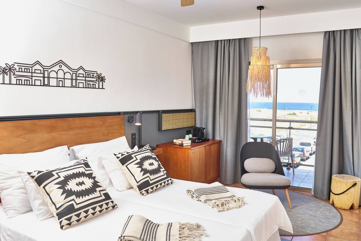 A hotel room with a bed, a desk with a lamp, a chair, a small table, and a view of the beach.
