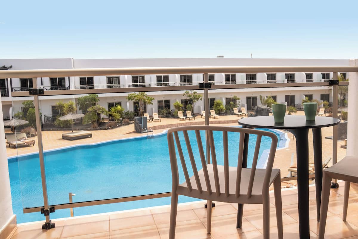 A balcony with a view of a swimming pool, lounge chairs, and a table with two green cups.