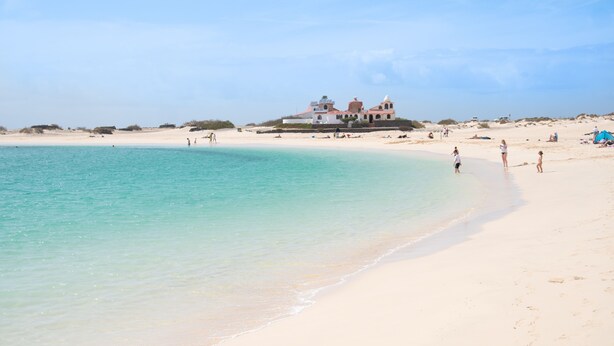 A beach with a large building in the background. - Coral Cotillo Beach (La Oliva)