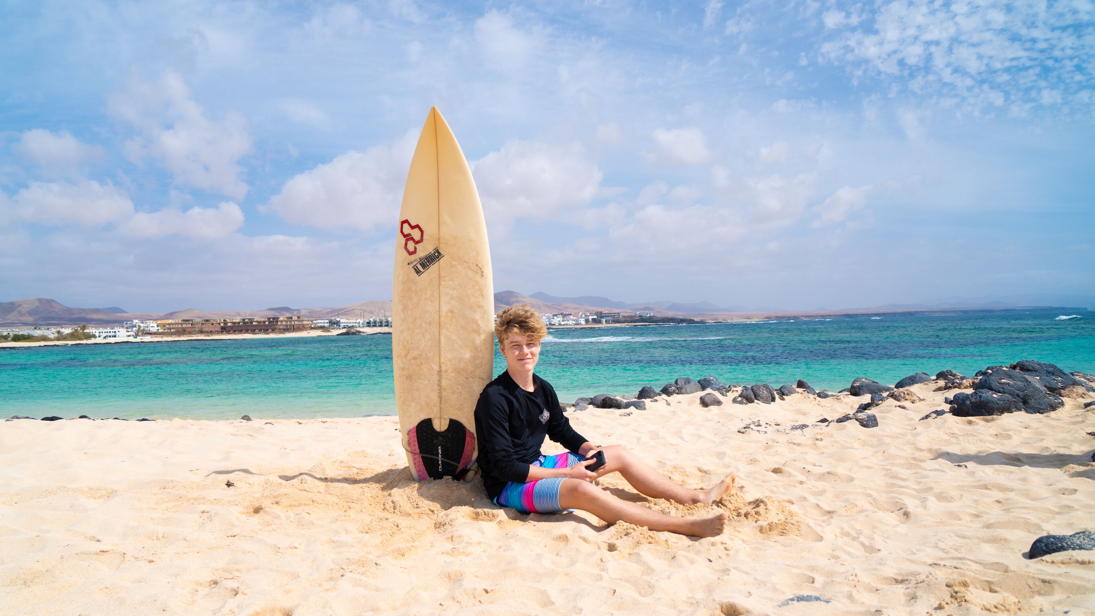 A person sitting on a sandy beach with a surfboard leaning against them.