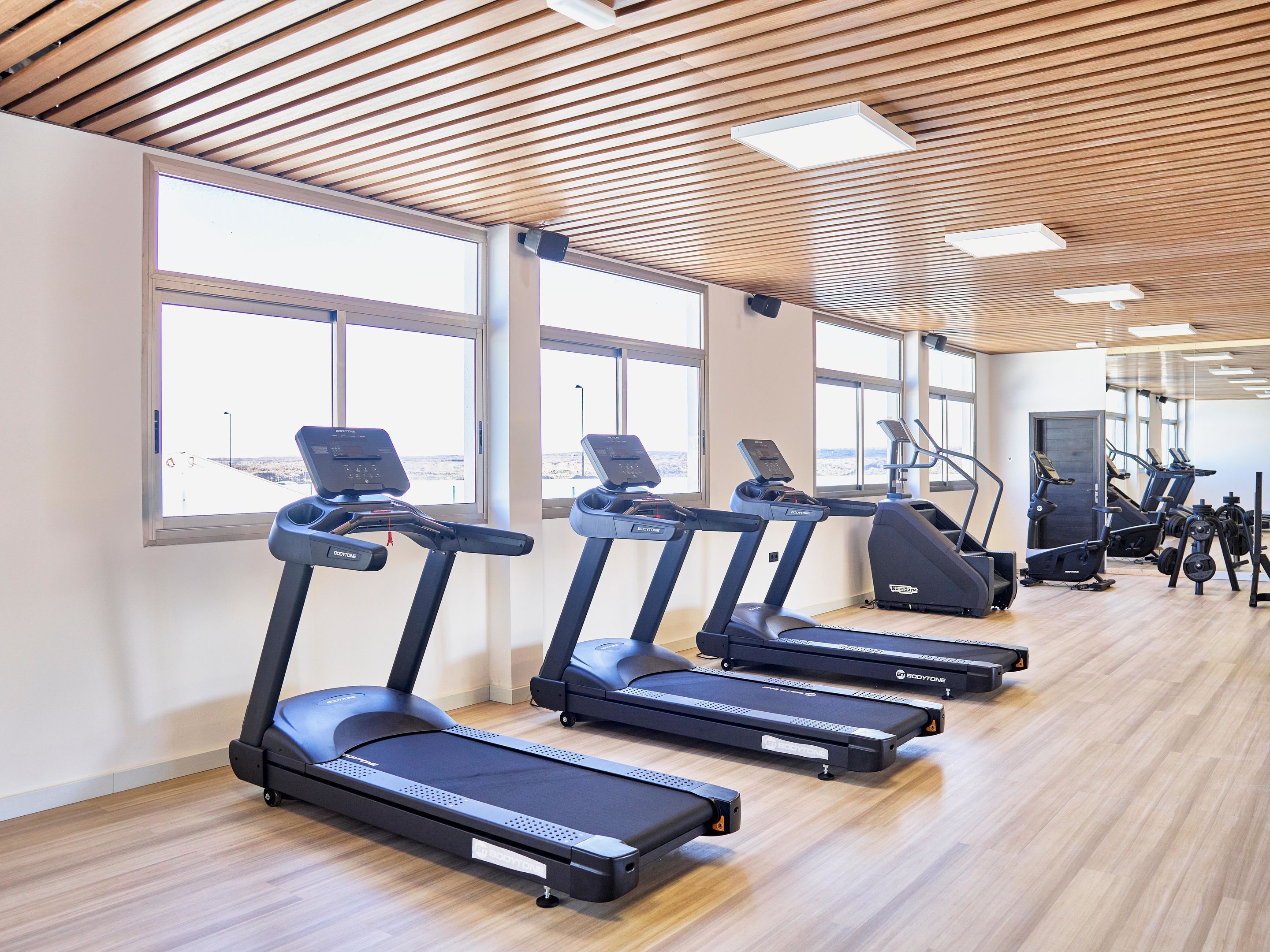 Un gimnasio con cintas de correr, bicicletas estáticas y máquinas de pesas, ventanas amplias y techo de madera.