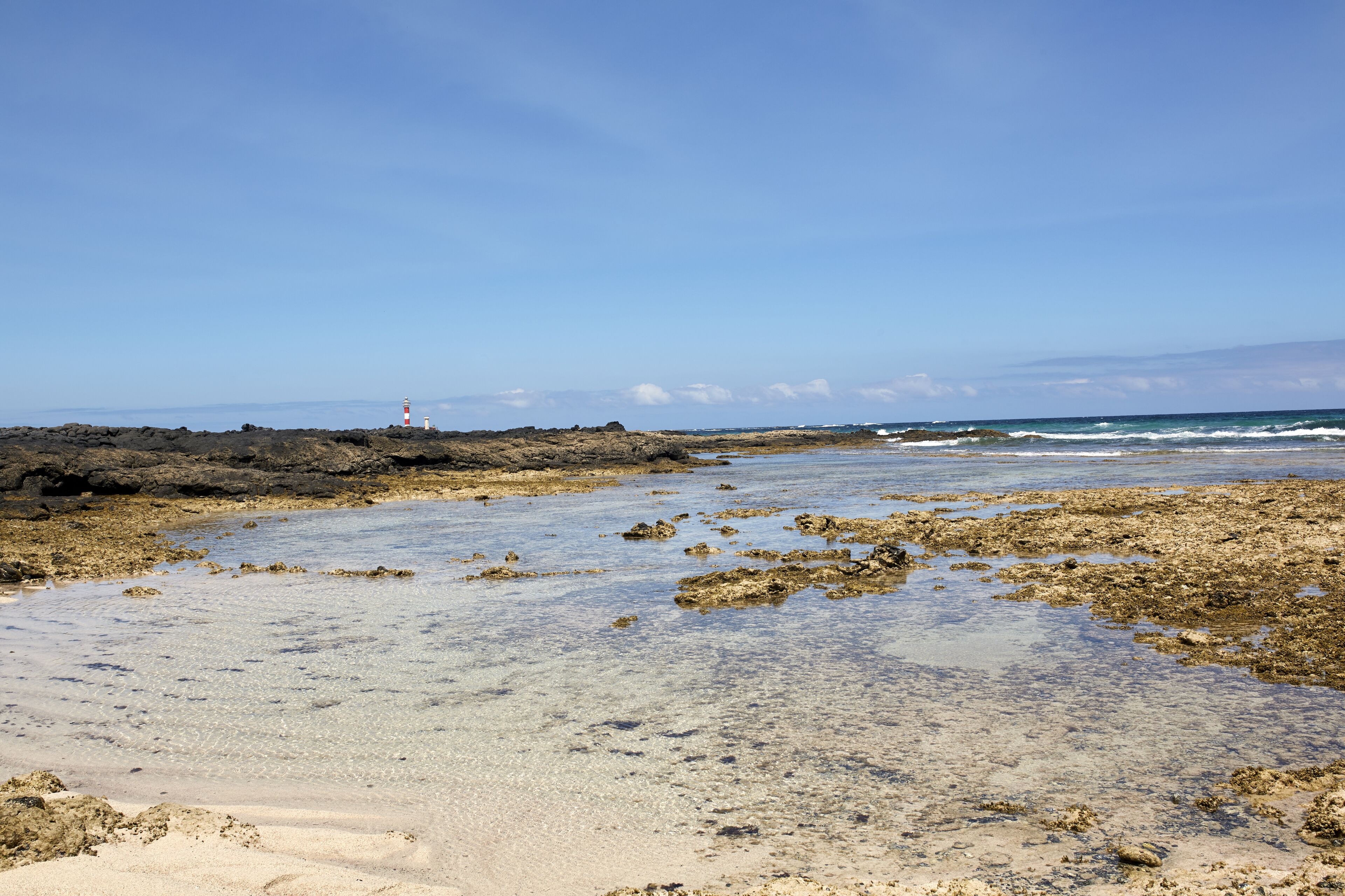 Un paysage côtier caractérisé par un terrain rocheux, des eaux claires et peu profondes, et un phare situé au loin.