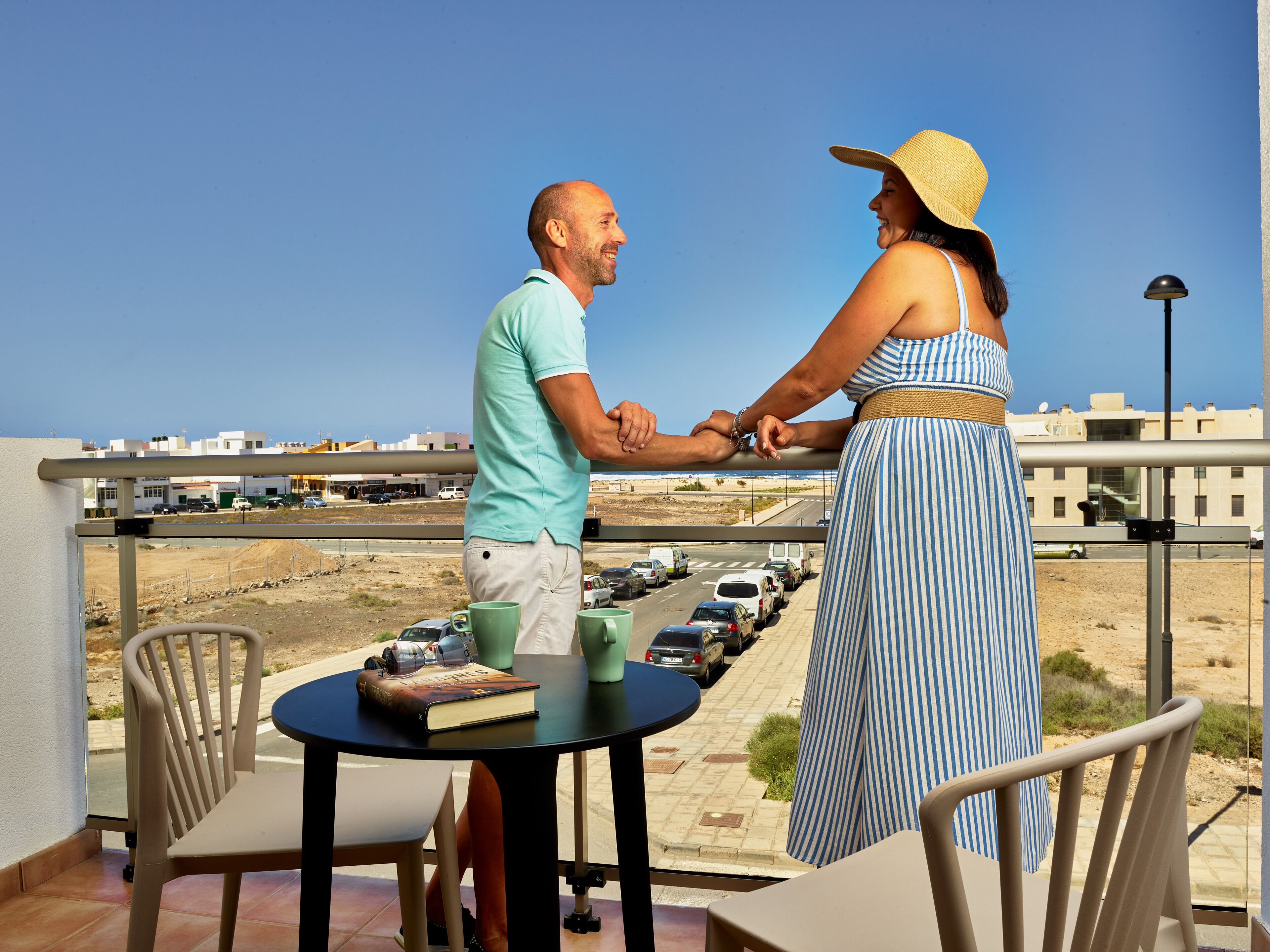 A couple holding hands on a balcony overlooking a parking lot and buildings.