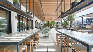 An outdoor dining area with long tables and wooden stools, surrounded by potted plants and a wooden ceiling structure. - Coral Cotillo Beach (La Oliva)