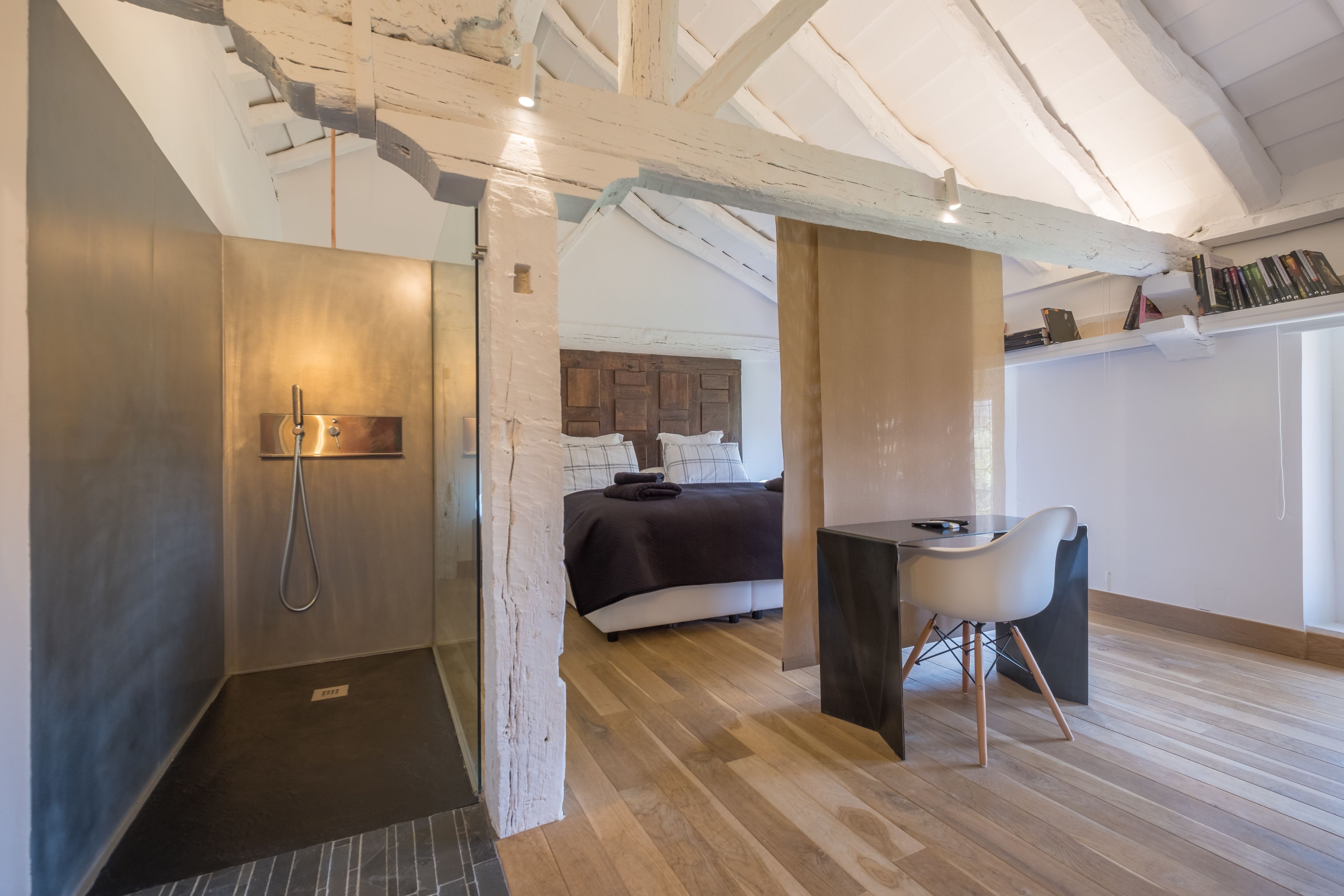 A modern bedroom with a wooden floor, a bed with a dark headboard, a desk with a chair, and a shower area.