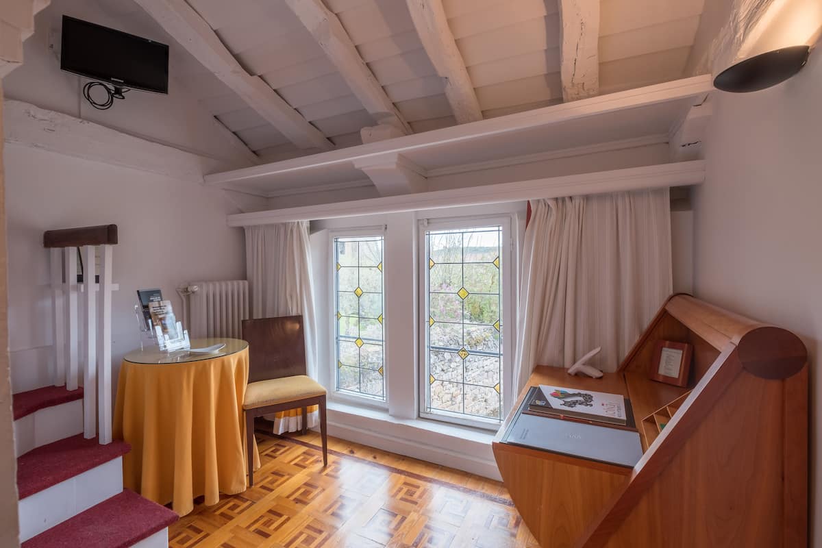 Een kamer met een houten trap, een ronde tafel met een geel tafelkleed, een stoel en een houten bank. Er zijn twee ramen met gordijnen, een radiator en een aan de muur bevestigde televisie.