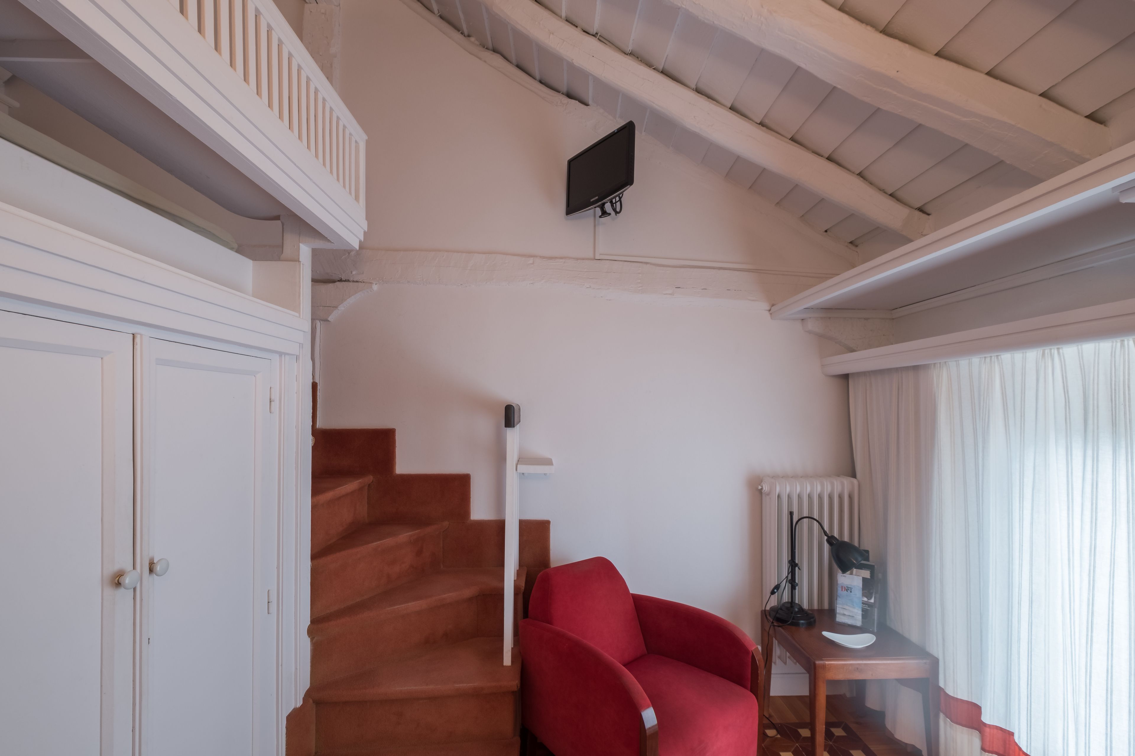 Una escalera conduce a un balcón, un sillón rojo, un escritorio con una lámpara y un televisor fijado a la pared.