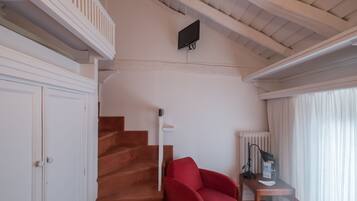 Una escalera conduce a un balcón, un sillón rojo, un escritorio con una lámpara y un televisor fijado a la pared.