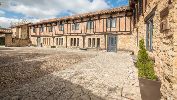 Une cour dallée de pierres, un bâtiment à ossature en bois et aux murs en pierre, et un petit bac à fleurs.