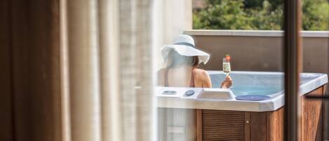 Une personne se détend dans un bain à remous, un verre à la main, entourée de lambris en bois et bénéficiant d’une vue sur la verdure.
