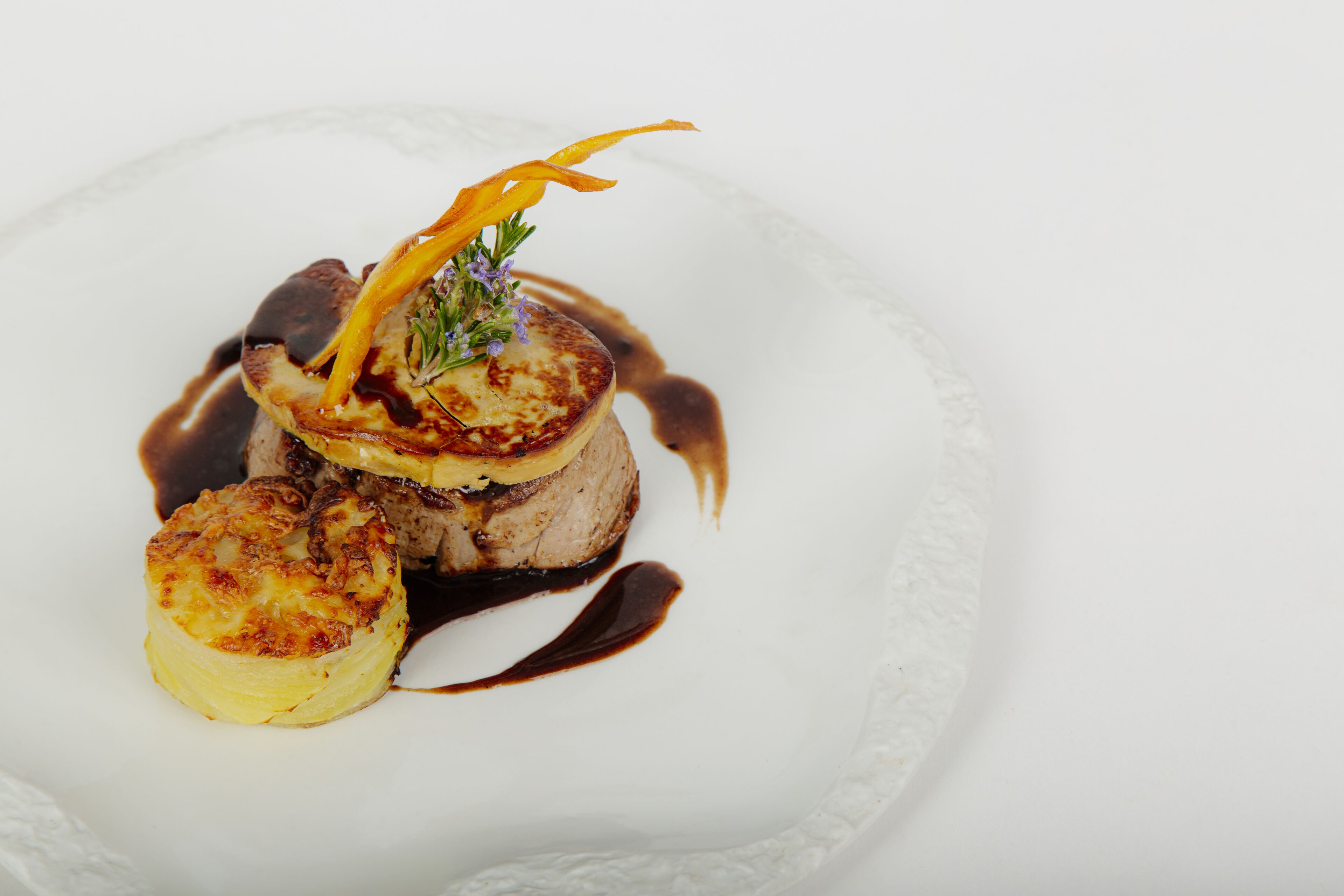 Um prato branco com uma porção de comida, incluindo um pedaço de carne, uma fatia de batata e um enfeite.