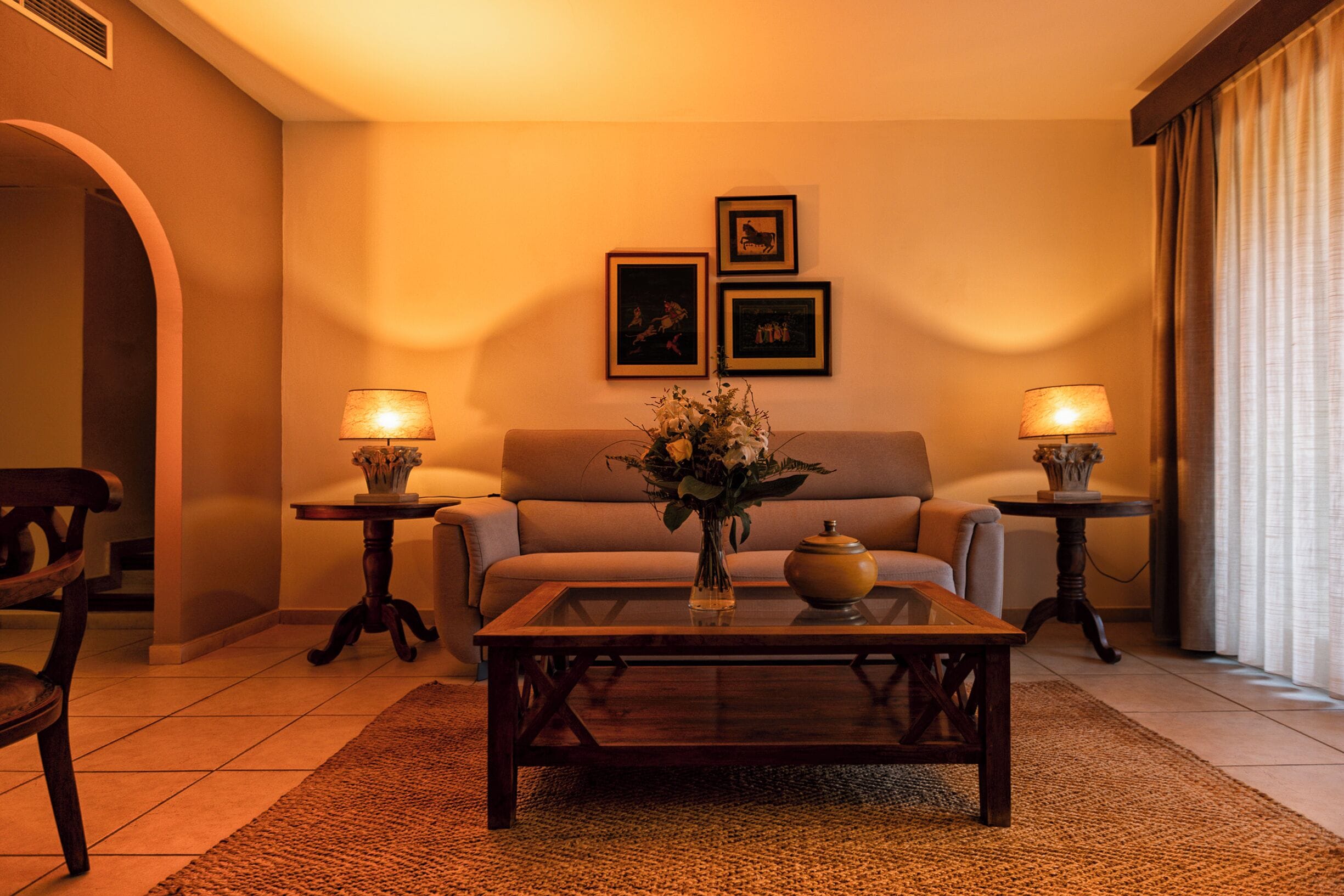 A cozy living room with a sofa, two side tables with lamps, a coffee table with a vase of flowers, and framed pictures on the wall.