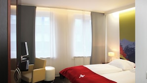 Habitación de hotel con cama, escritorio, silla, televisión y ventana con cortinas.