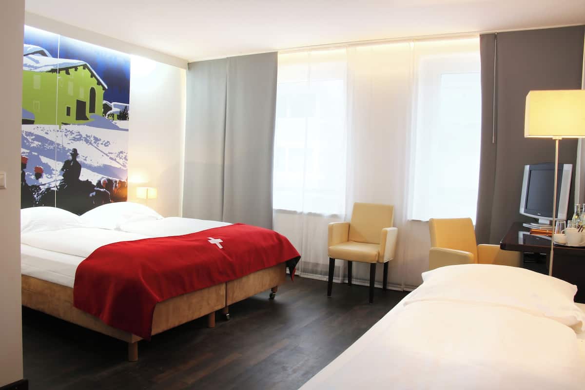 A hotel room with a bed, two chairs, a desk with a lamp, and a television.