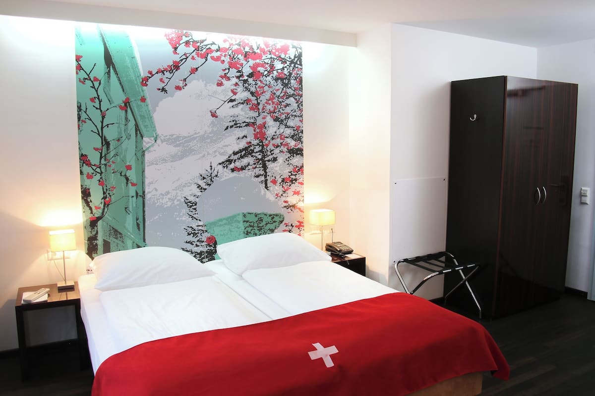 A hotel room with a bed, a nightstand, a lamp, a wardrobe, and a wall with a floral tapestry.