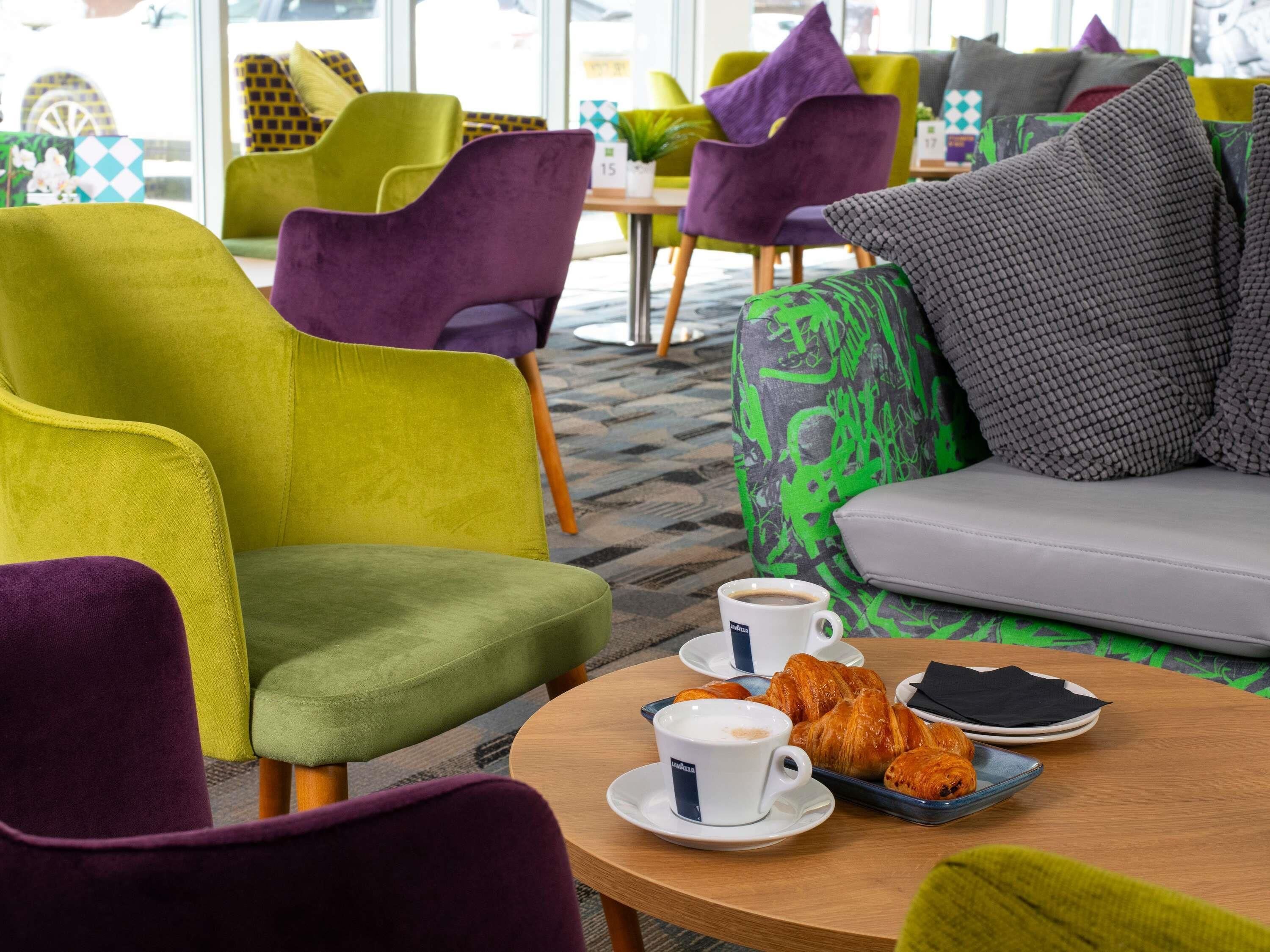 Ein moderner Lounge-Bereich mit lebendigen Sesseln, einem runden Tisch mit einer Tasse Kaffee und Gebäck, und Blick durch große Fenster nach draußen.