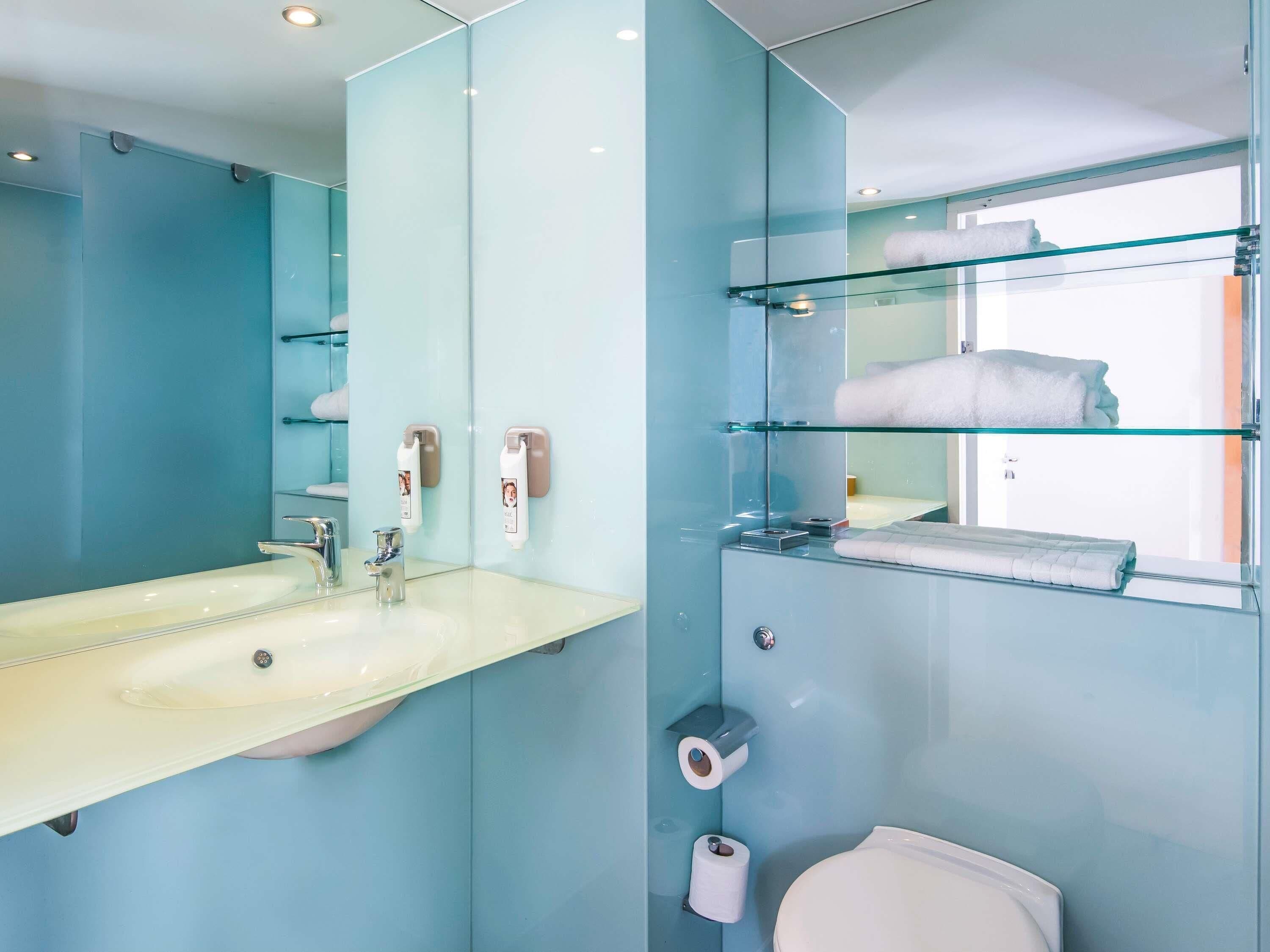 A modern bathroom with a large mirror, a sink, a toilet, and glass shelves for towels.
