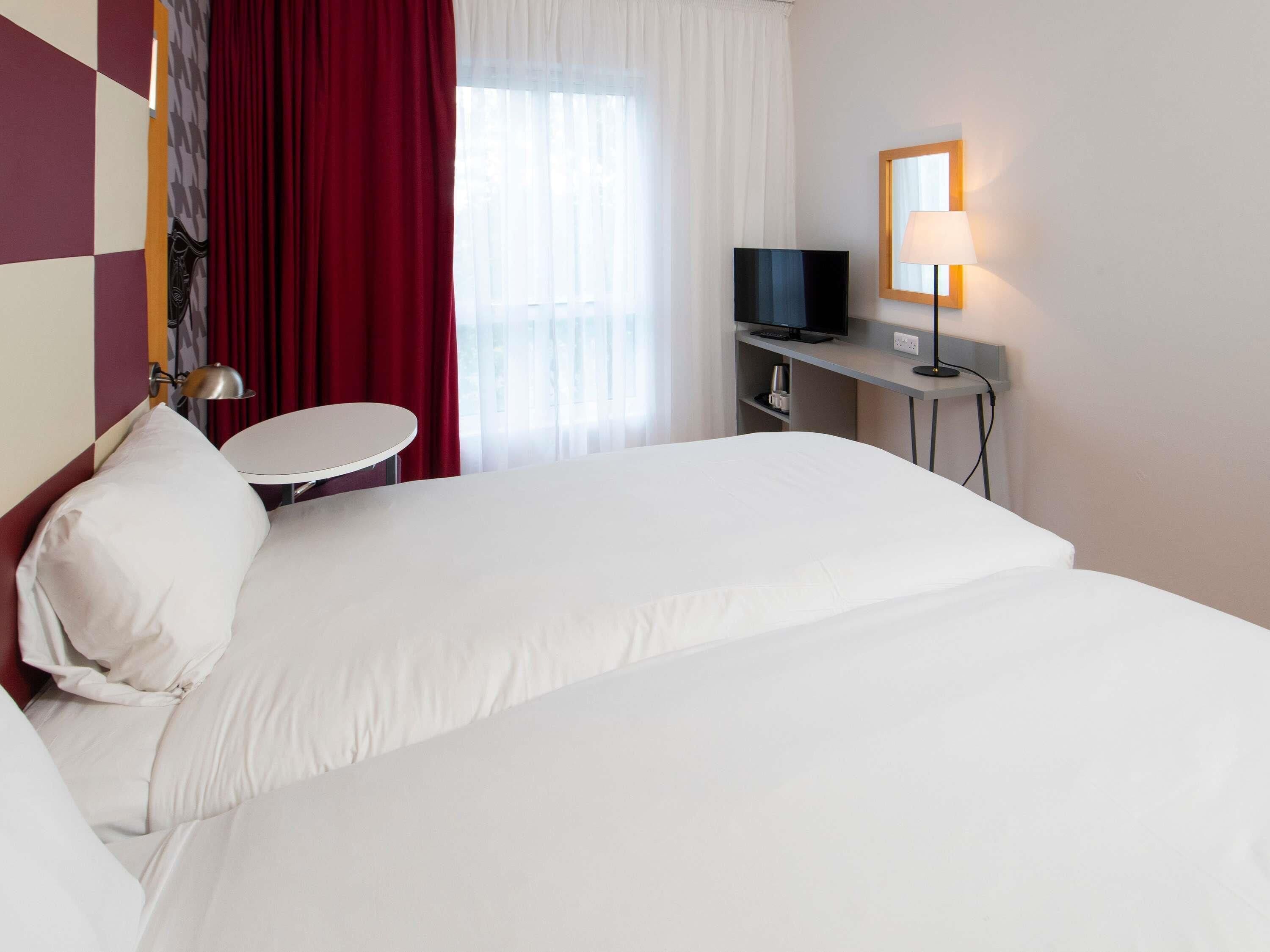 A hotel room with a bed, a desk with a TV, a lamp, and a window with curtains.