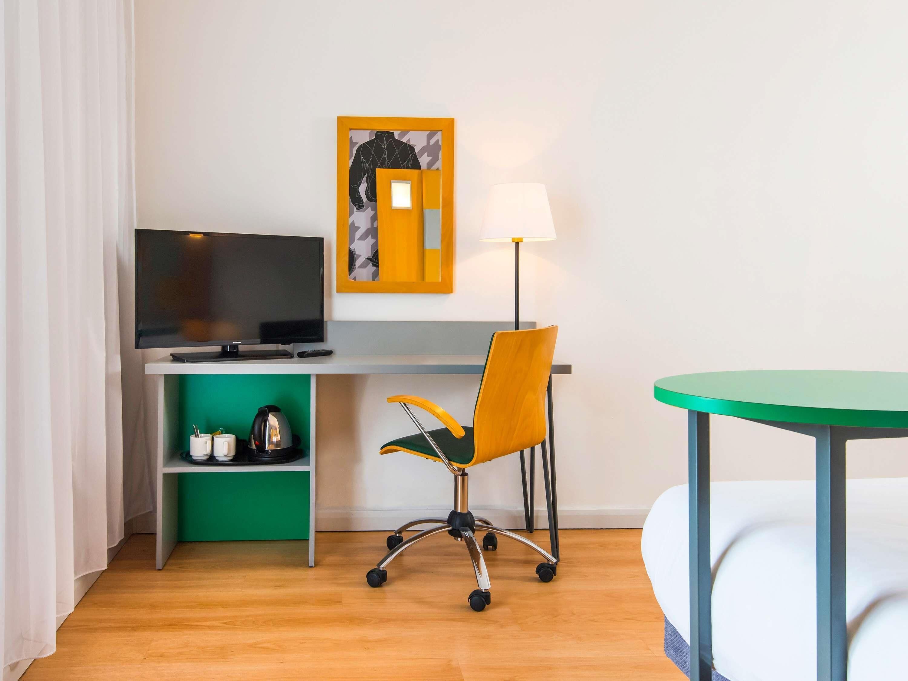 A hotel room with a desk, chair, TV, lamp, and a bed.
